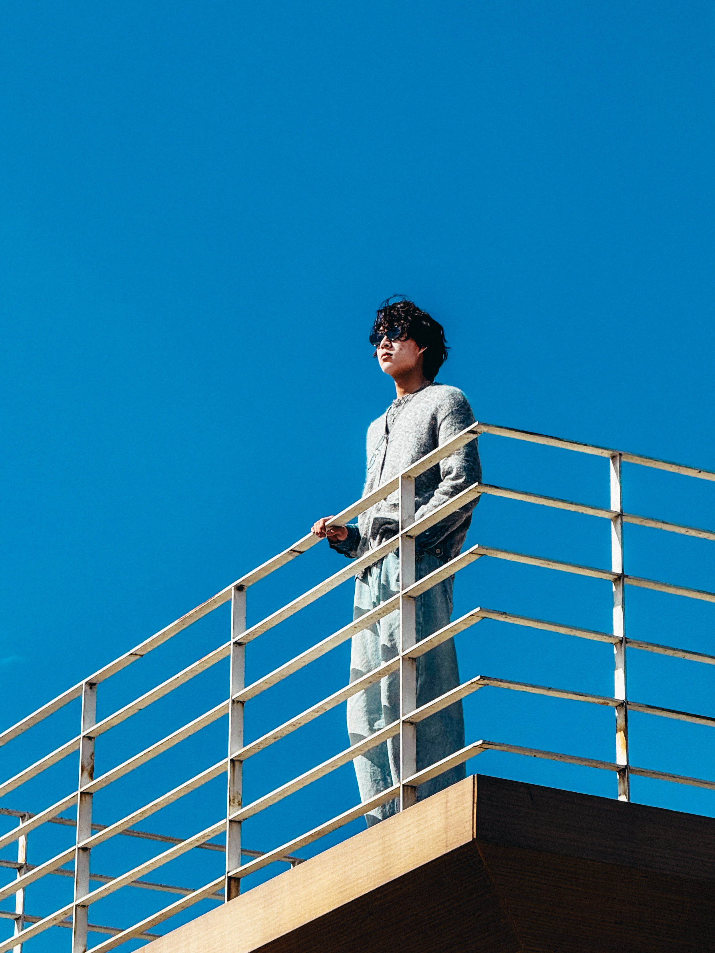 A person with dark curly hair and sunglasses standing on a balcony with a white railing, against a clear blue sky.