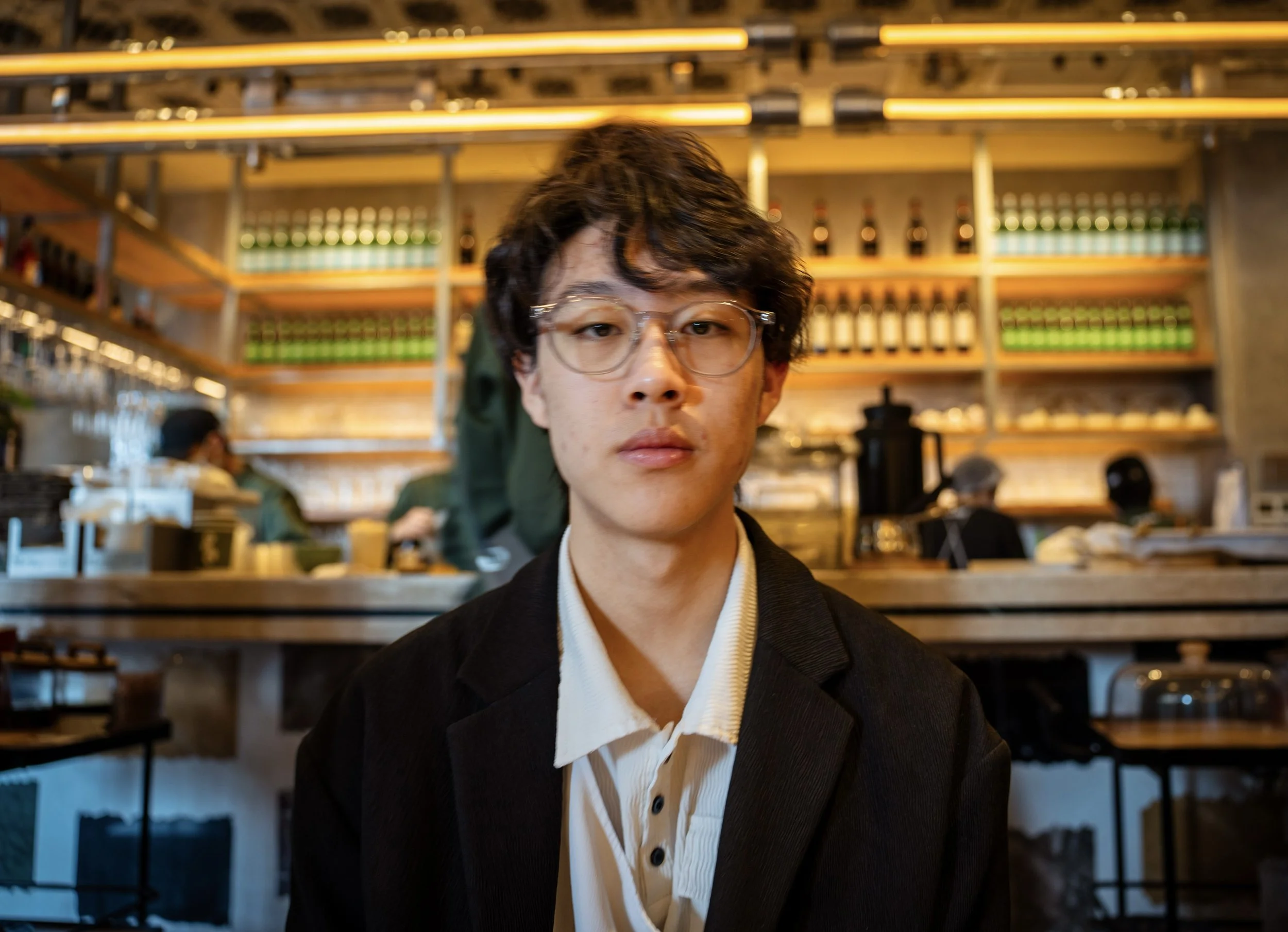 A young man with glasses and wavy dark hair sitting in a café or restaurant, with shelves of wine bottles and cooking tools behind him, and people working in the background.