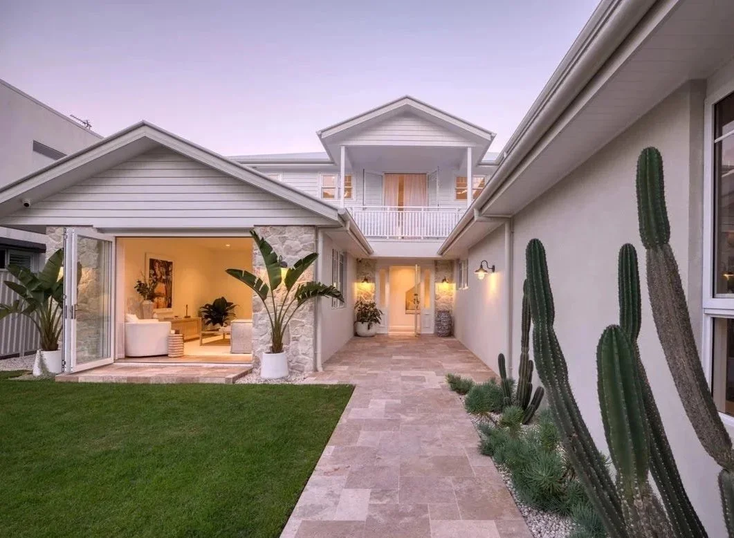 Modern house renovation with a patio, green grass, large cacti, and potted plants, with indoor living room visible through open doors.