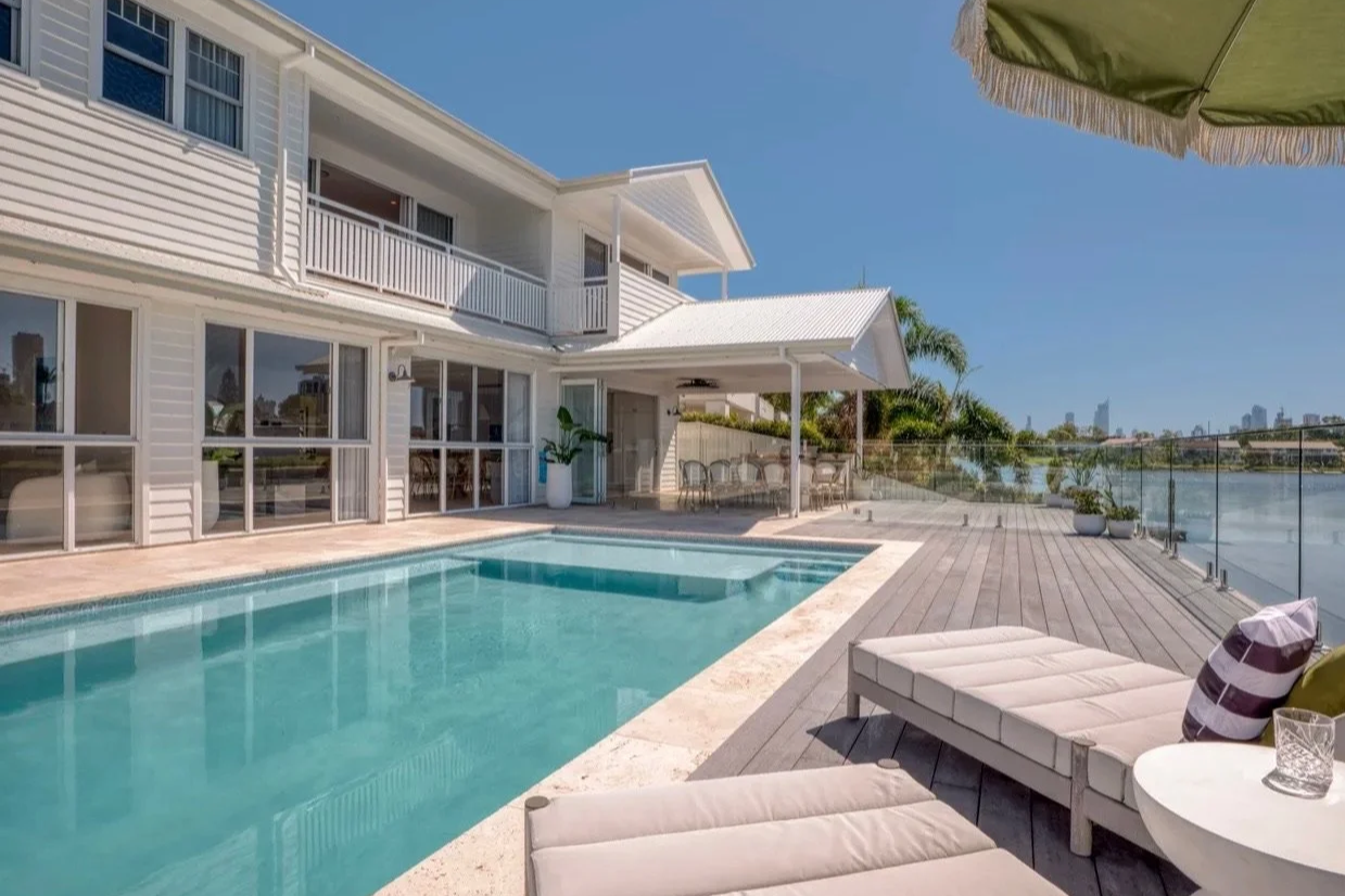 A backyard with a swimming pool next to a modern white house on a clear, sunny day. There are lounge chairs and a patio dining area with trees and city skyline in the distance.