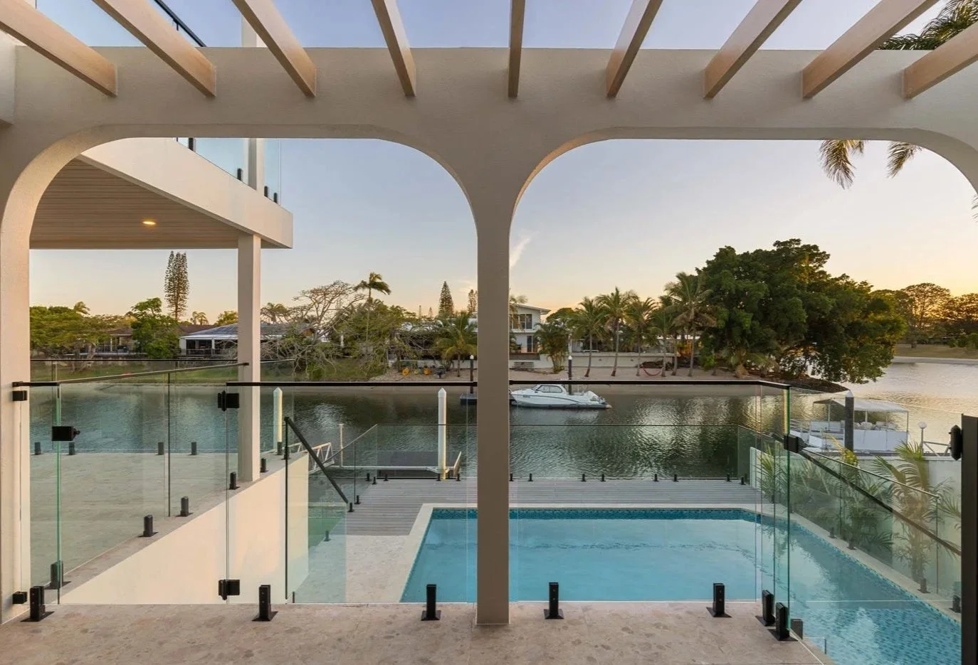 View from a balcony overlooking a private pool, waterway, and lush trees with a boat docked in the water during sunset.