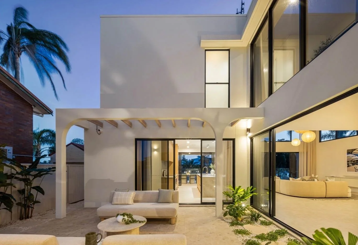 Modern house with outdoor seating area, large glass windows, and indoor living room visible inside, during evening time.