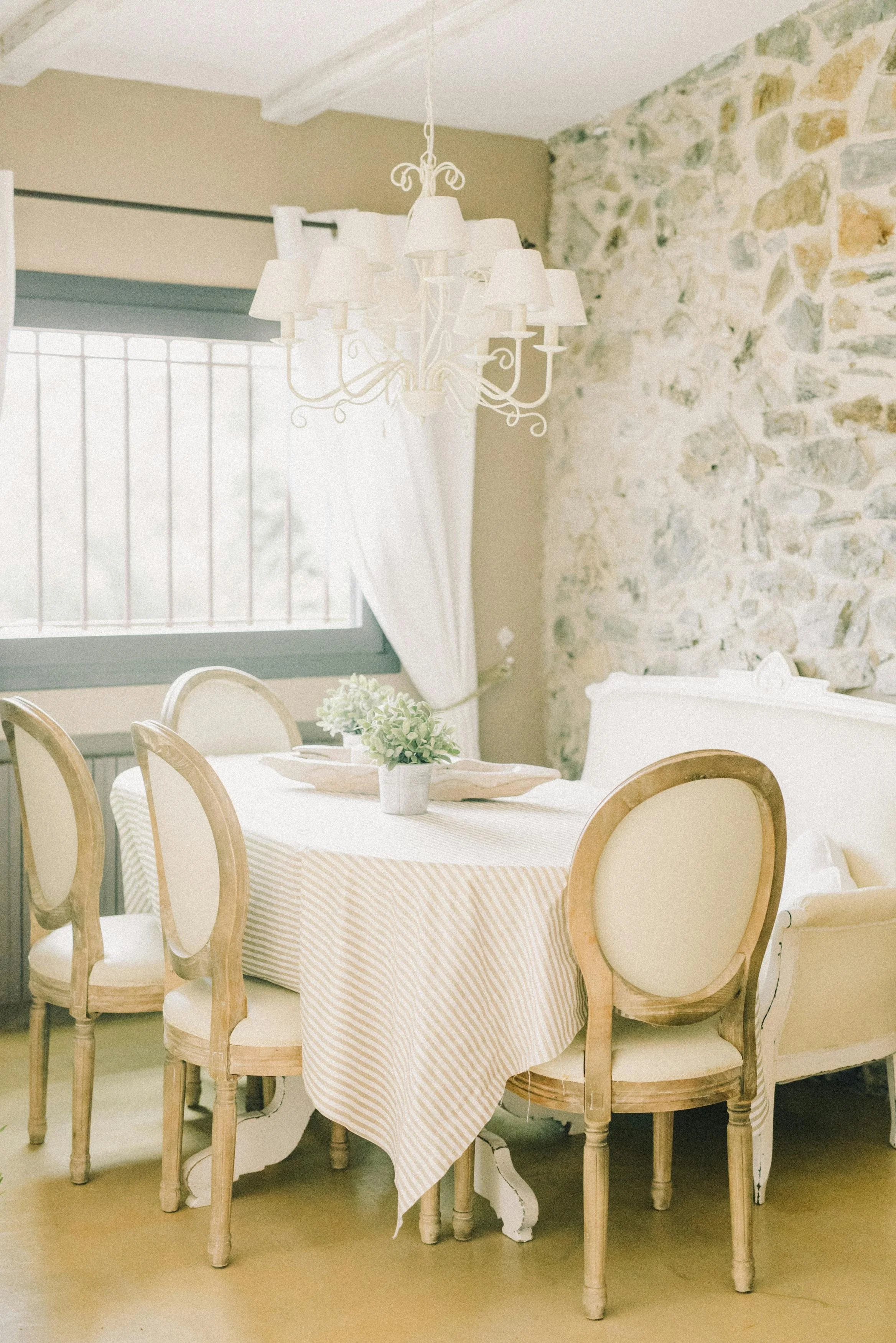 A dining room with a vintage style. It features a round table with a striped tablecloth, surrounded by wooden chairs with cream upholstery, a white chandelier hanging above, a window with curtains, and a stone wall.