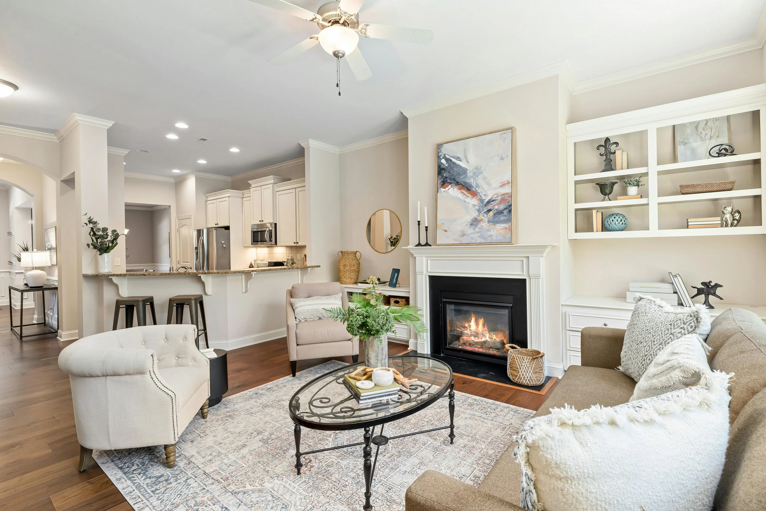 Living room with a fireplace, beige sofa, armchair, glass coffee table with decorative items, built-in shelves, and an open kitchen with white cabinets and stainless steel appliances.