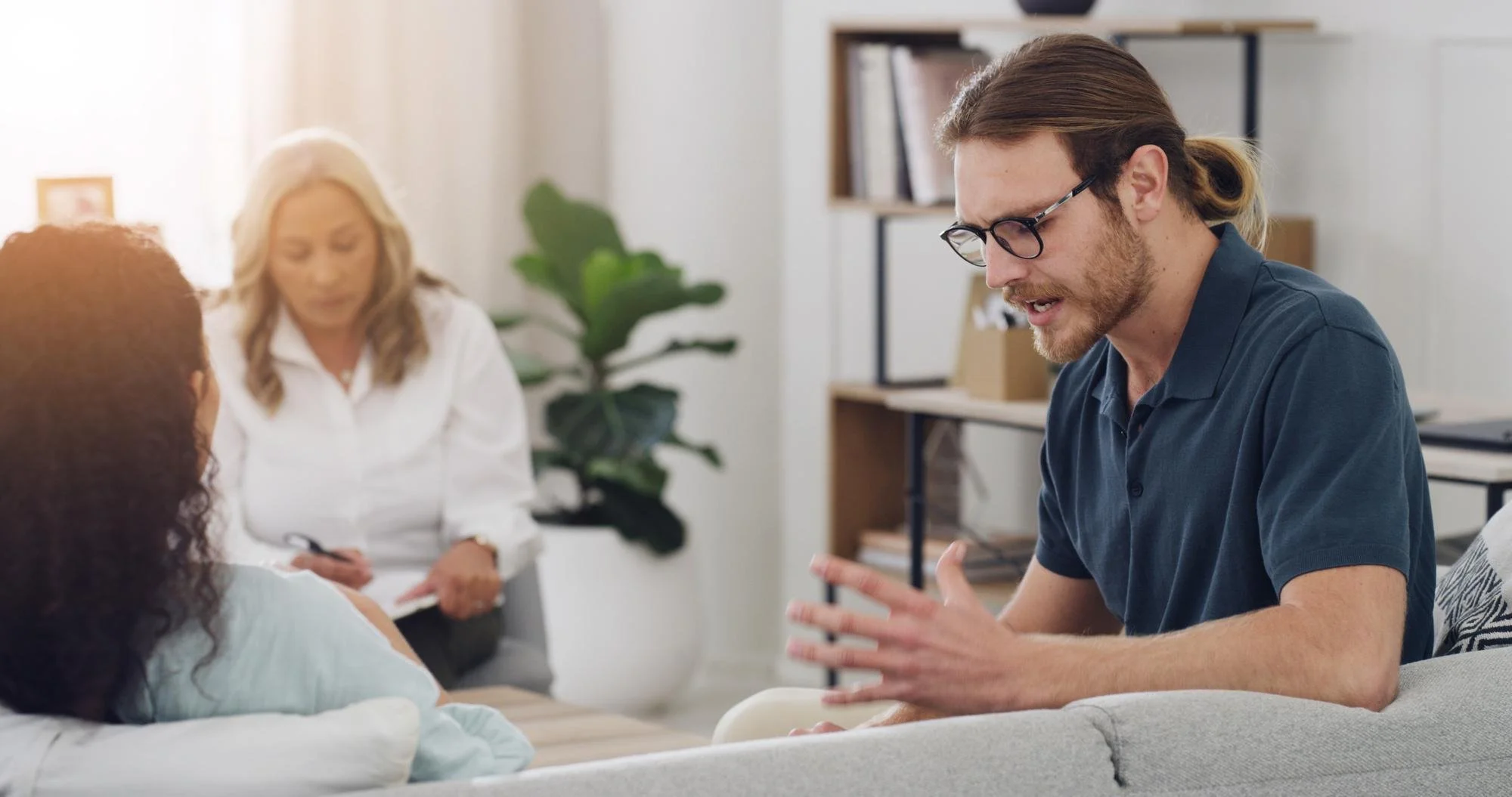 A man with glasses and long hair tied back, sitting on a sofa, talking to a woman lying on the sofa. In the background, a woman with gray hair taking notes, with a plant and bookshelf in the room.