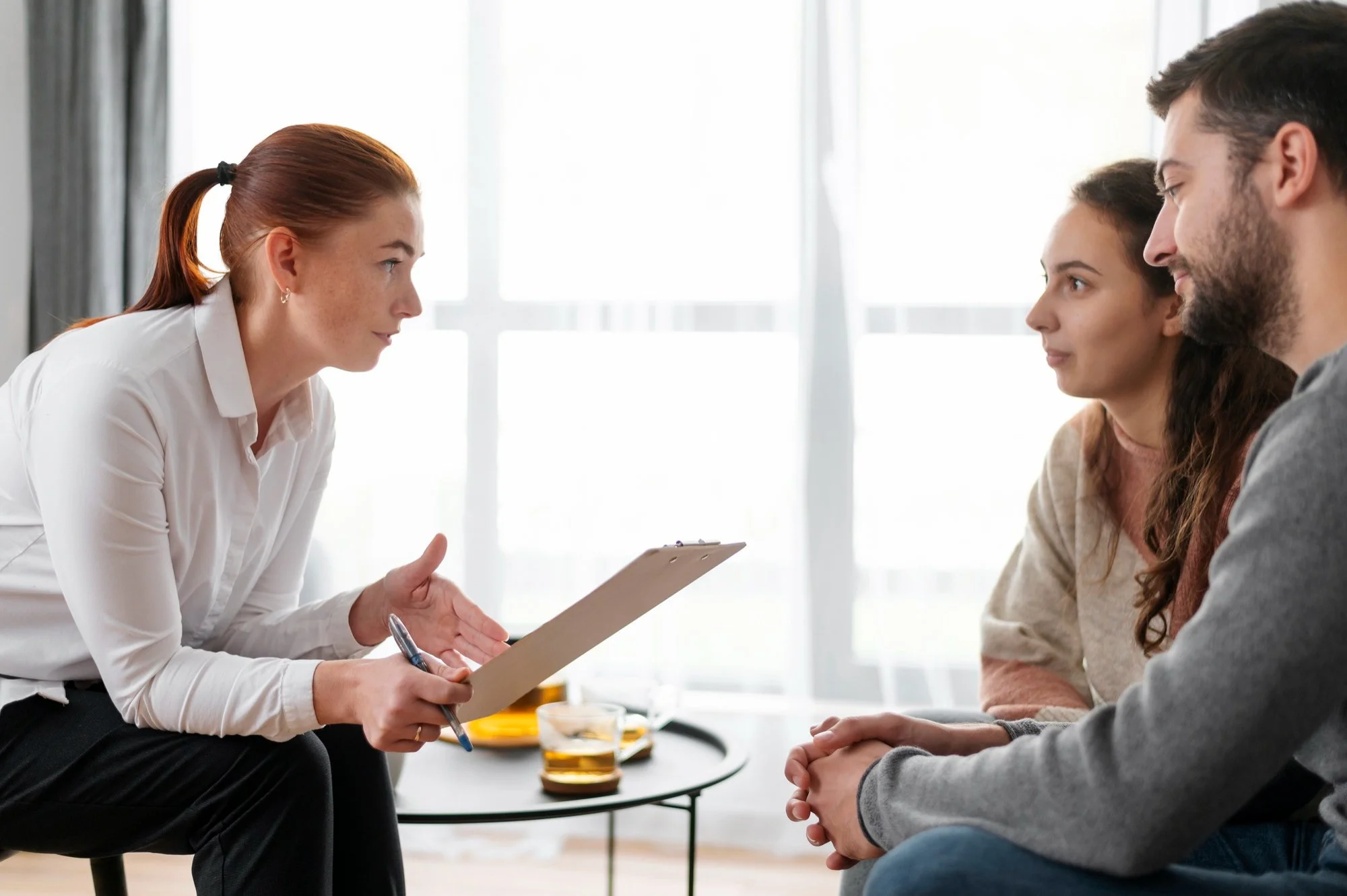 A woman therapist taking notes during a therapy session with a man and a woman patient sitting across from her in a bright room.