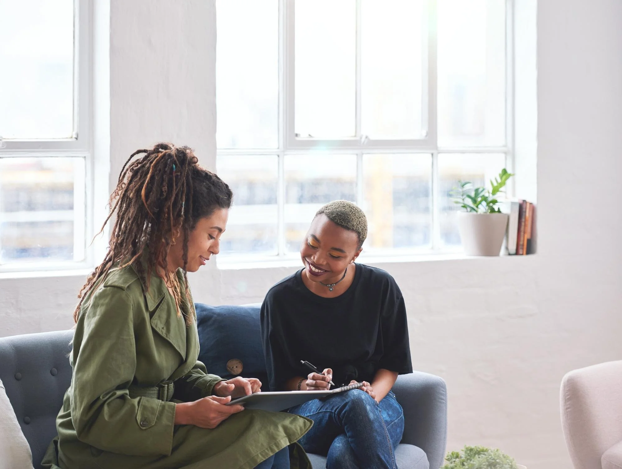 Two women sitting on a couch smiling and talking, one holding a tablet and the other holding a pen and notebook, in a bright room with large windows, plants, and books.