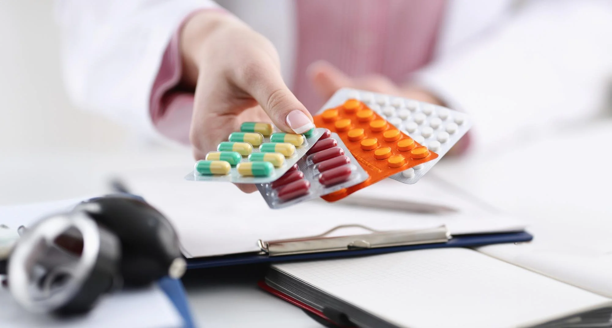 A hand holding three blister packs of medication, with various colorful pills and capsules, on a white medical or desk surface with a stethoscope, clipboard, and notebook in the background.