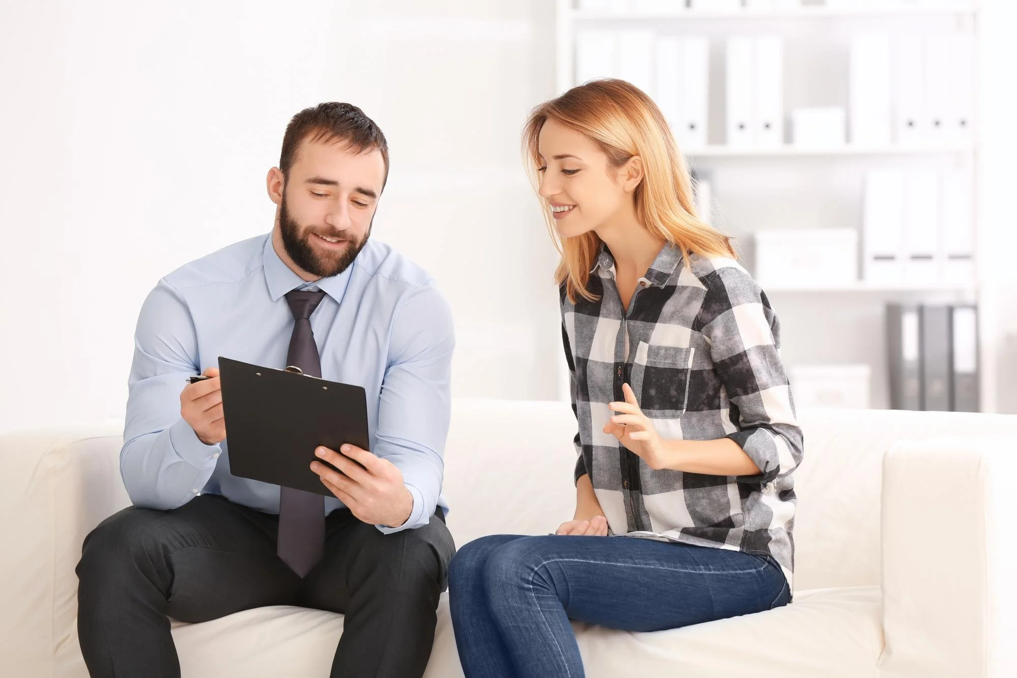 A man in a light blue shirt and dark tie holding a clipboard, smiling, as he talks to a woman with blonde hair in a checkered shirt, sitting on a white couch in a modern office space.