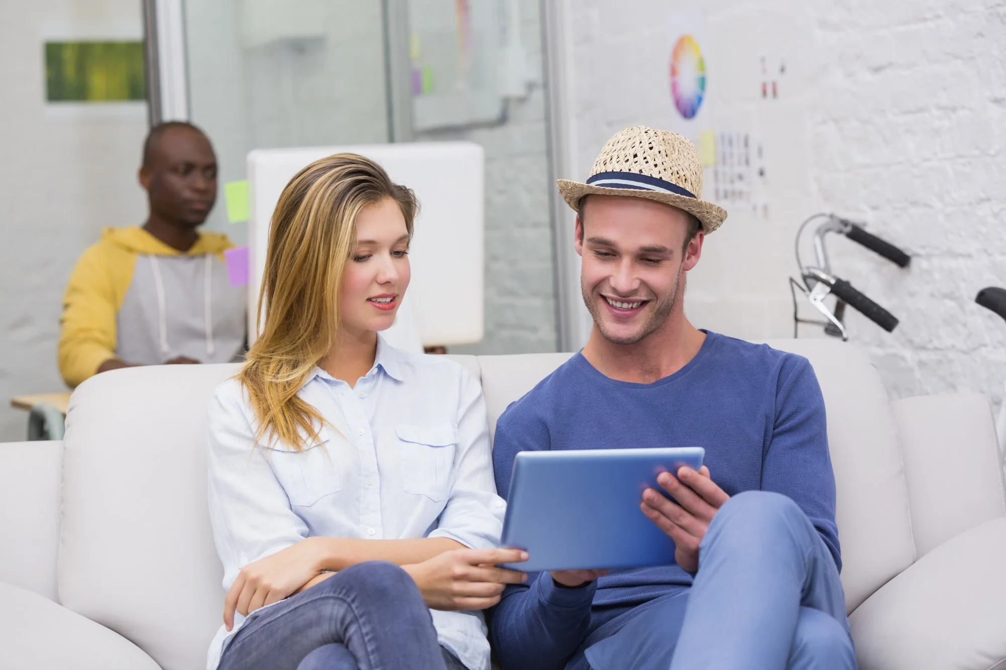 Two young adults, a man wearing a straw hat and a woman, sit on a white couch, looking at a tablet together and smiling. A man in a yellow hoodie works at a desk in the background, with colorful decor and a whiteboard.