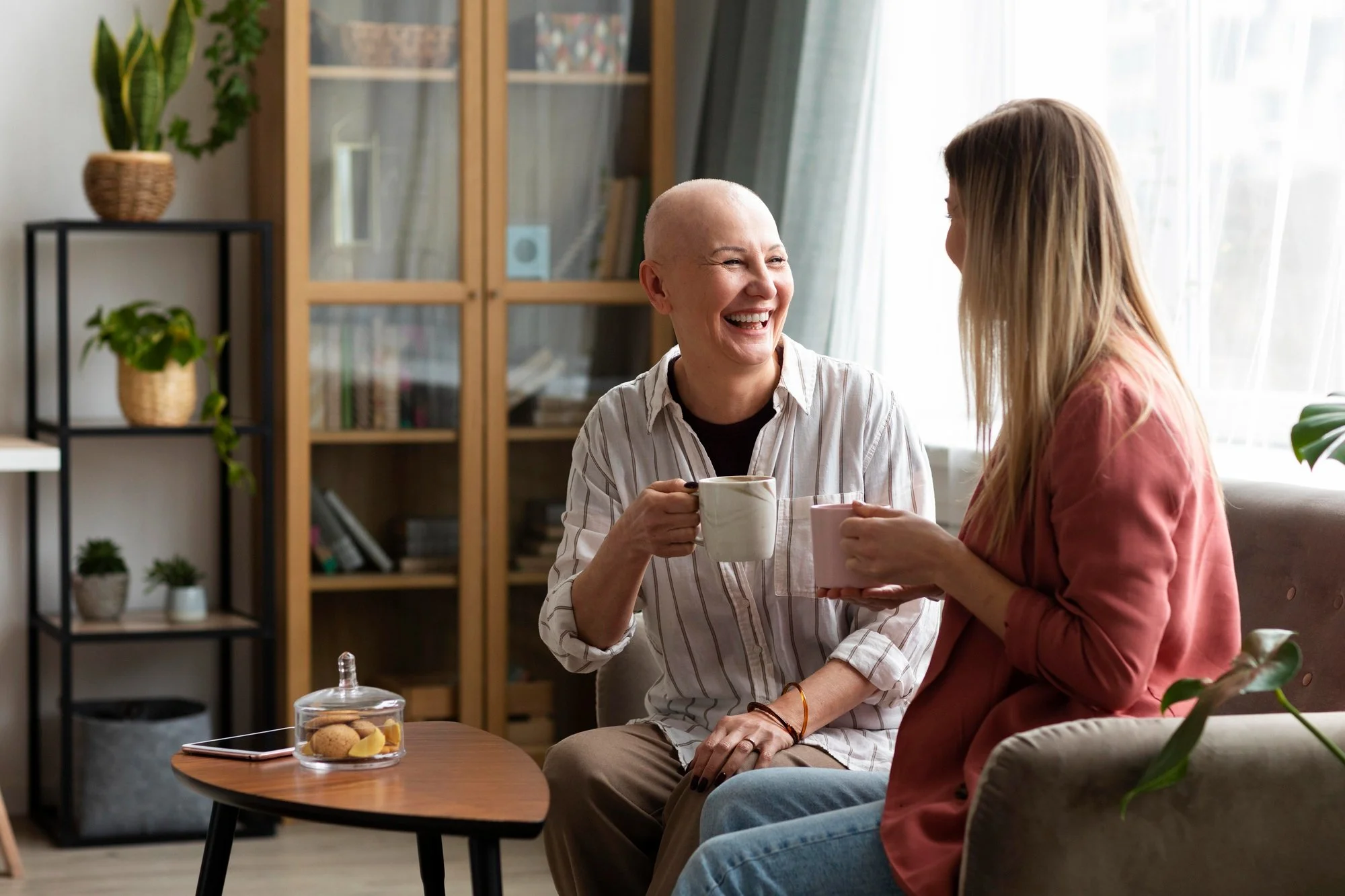 Two women sitting on a sofa, sharing a laugh and holding mugs in a cozy living room with bookshelves and houseplants.
