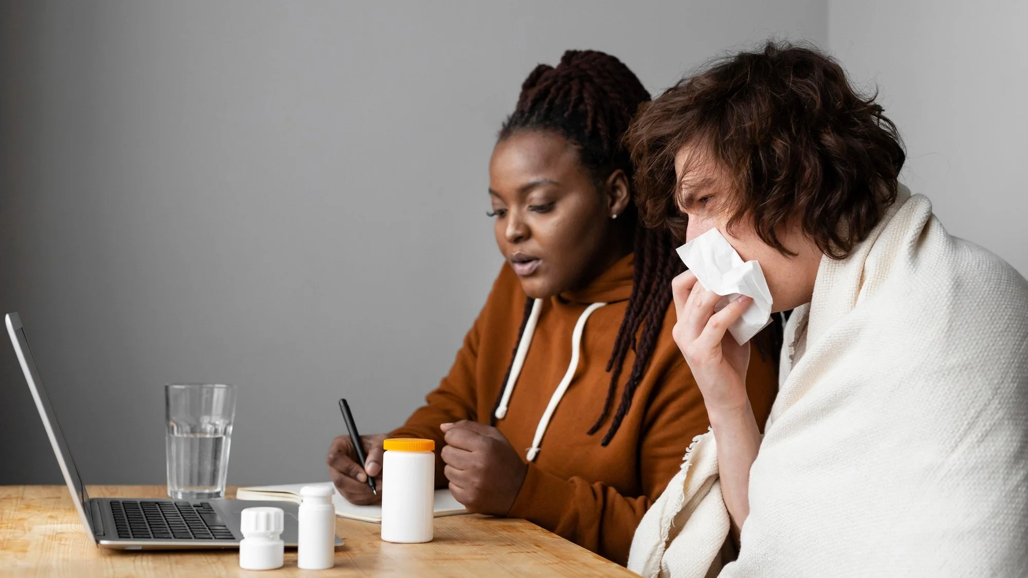 A woman with short brown hair wrapped in a white blanket, sneezing into a tissue, sitting next to a woman with long, dark, dreadlocked hair wearing a brown hoodie, who is writing on a notepad at a table with medicine bottles, a glass of water, a laptop, and a notebook.