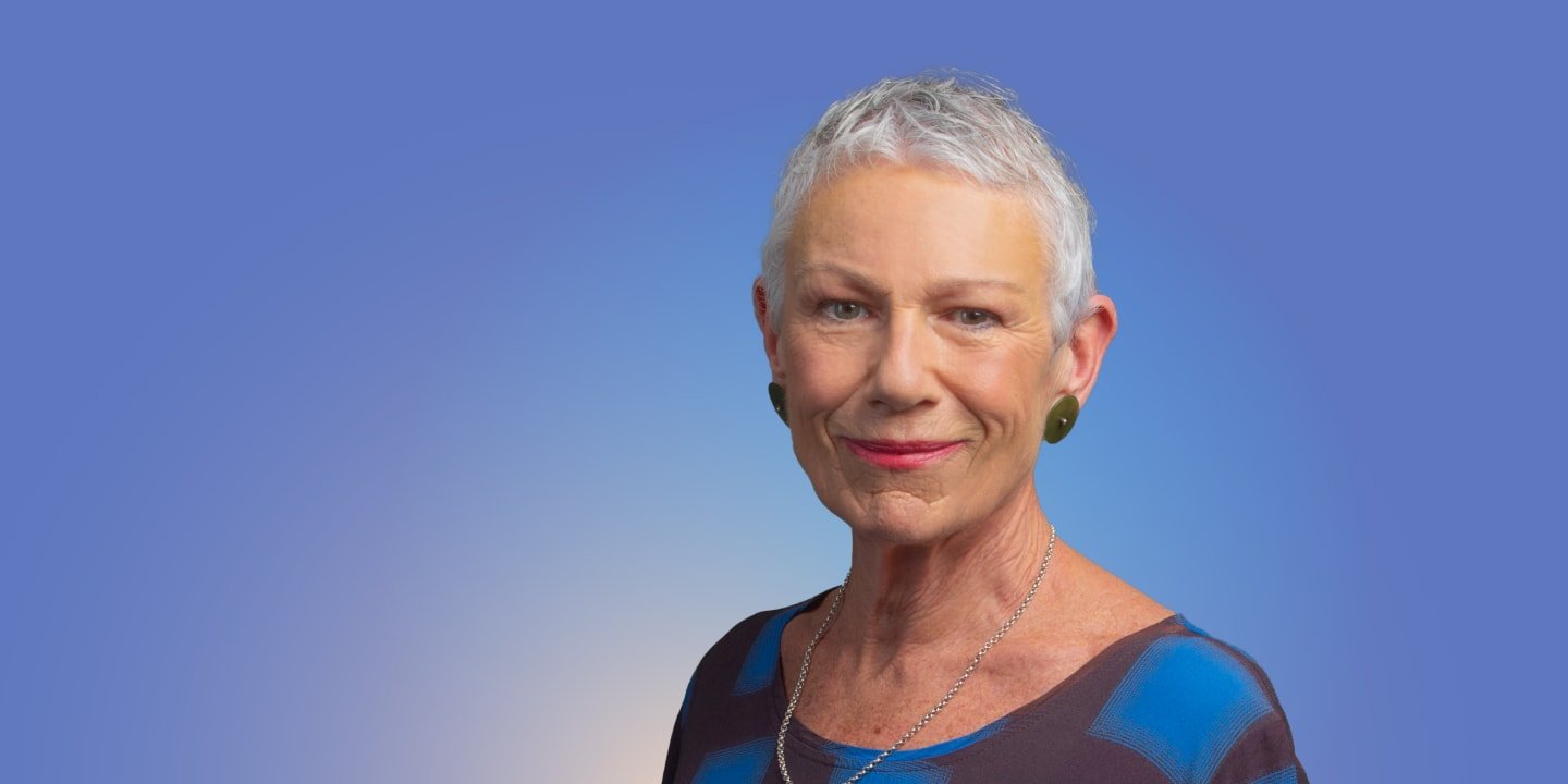 A woman, Kim Hill,  with short white hair, wearing a blue and black top and green earrings, stands against a blue gradient background.