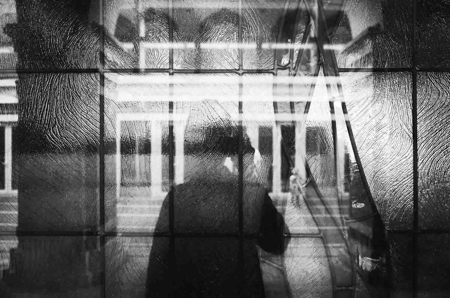 Black and white image of a person seen through a frosted glass window with a distorted reflection, creating a mysterious and abstract effect.