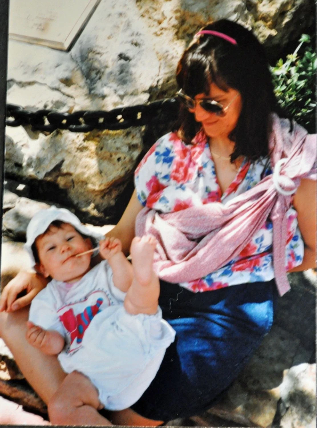Amanda Barusch wearing sunglasses and her baby in a hat sit outdoors. The baby holds a twig and rests feet on Amanda.