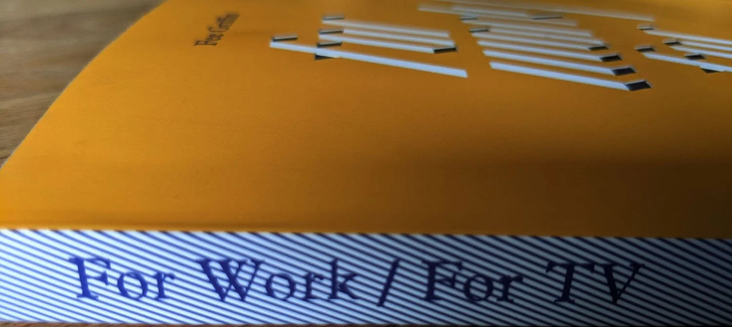 Close-up of a yellow book with text partially visible on the front and spine. The spine reads "For Work / For TV." The background is a wooden surface.