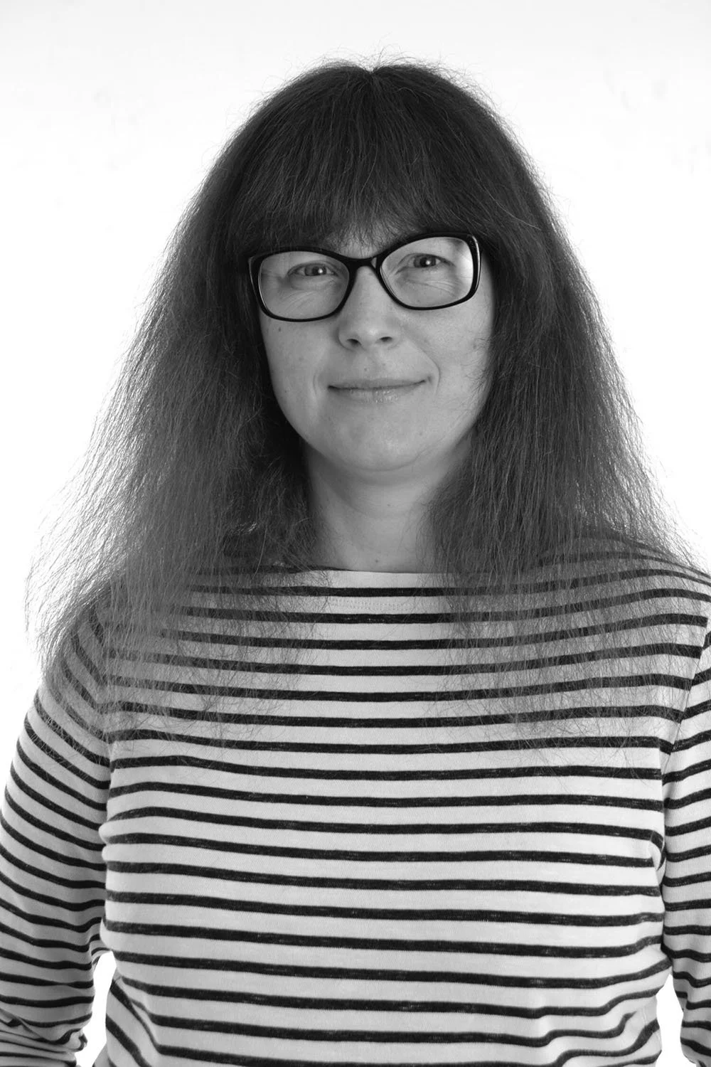 This is a black-and-white photo of Fee Griffin, who has long hair and glasses, is wearing a striped shirt, and is smiling at the camera.
