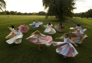 Twelve twirling women on a green field. Their colorful dresses make them look like pinwheels.