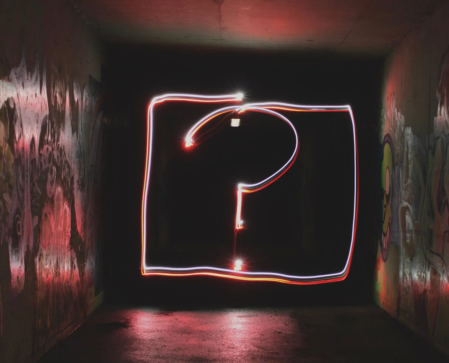A neon-red question mark outlined with white light on a dark background, within a graffiti-decorated tunnel.