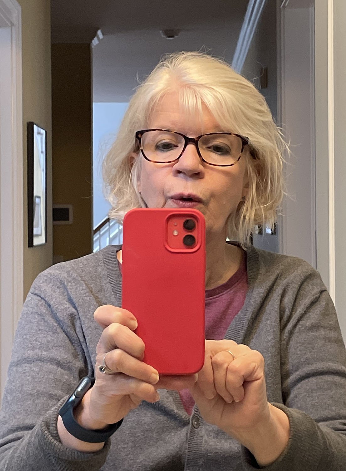 Katy Betts Adams - a woman with short, white hair and glasses, takes a selfie in a hallway using a red smartphone. She is wearing a gray sweater and a watch.