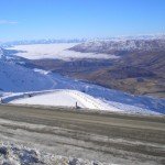 Mountainous landscape with partially snow-covered slopes and a clear blue sky. A winding road is seen in the foreground.
