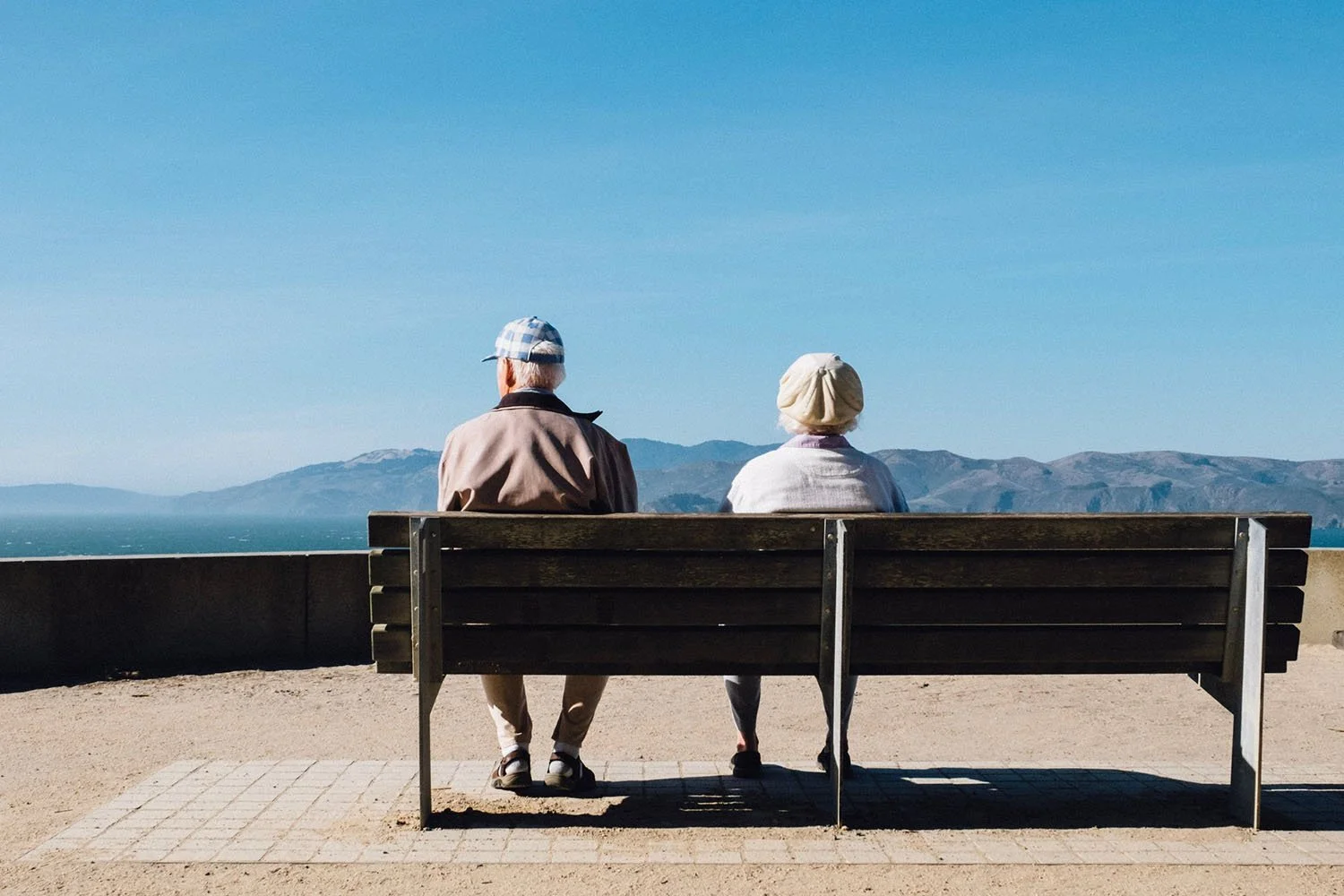 Two elderly people sit on a bench overlooking a vast landscape with mountains and a clear sky in the background.