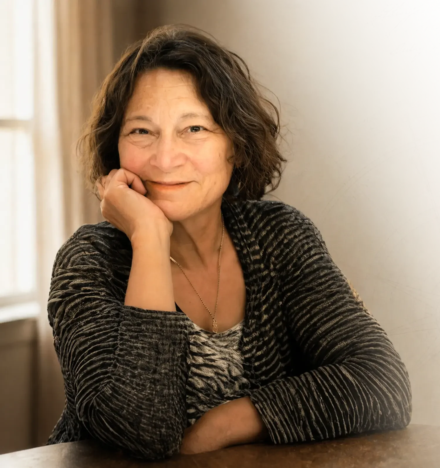 A woman with curly dark hair sitting at a table, resting her chin on her hand, smiling at the camera.