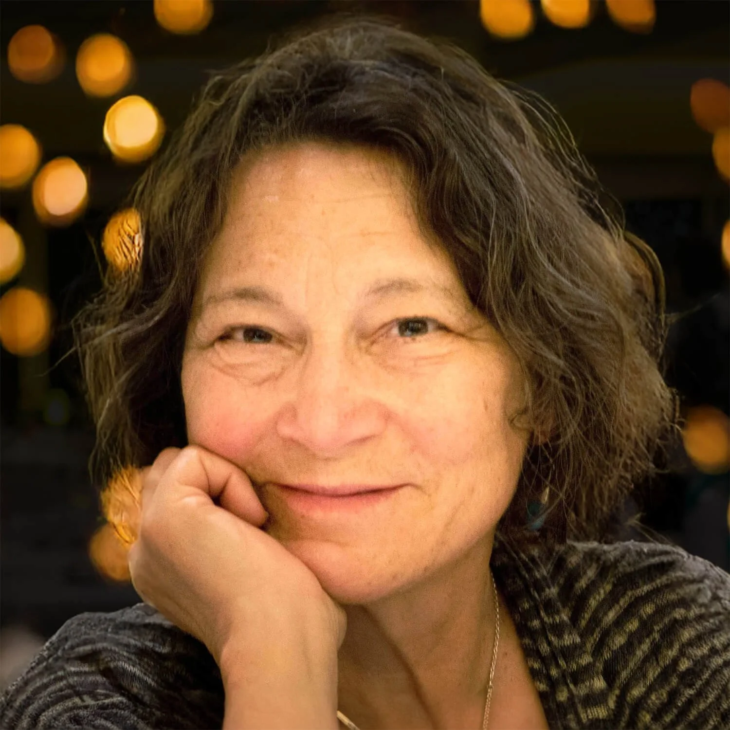 Close-up of a middle-aged woman with short wavy hair, smiling gently, with her hand resting on her chin, against a background with out-of-focus yellow lights.