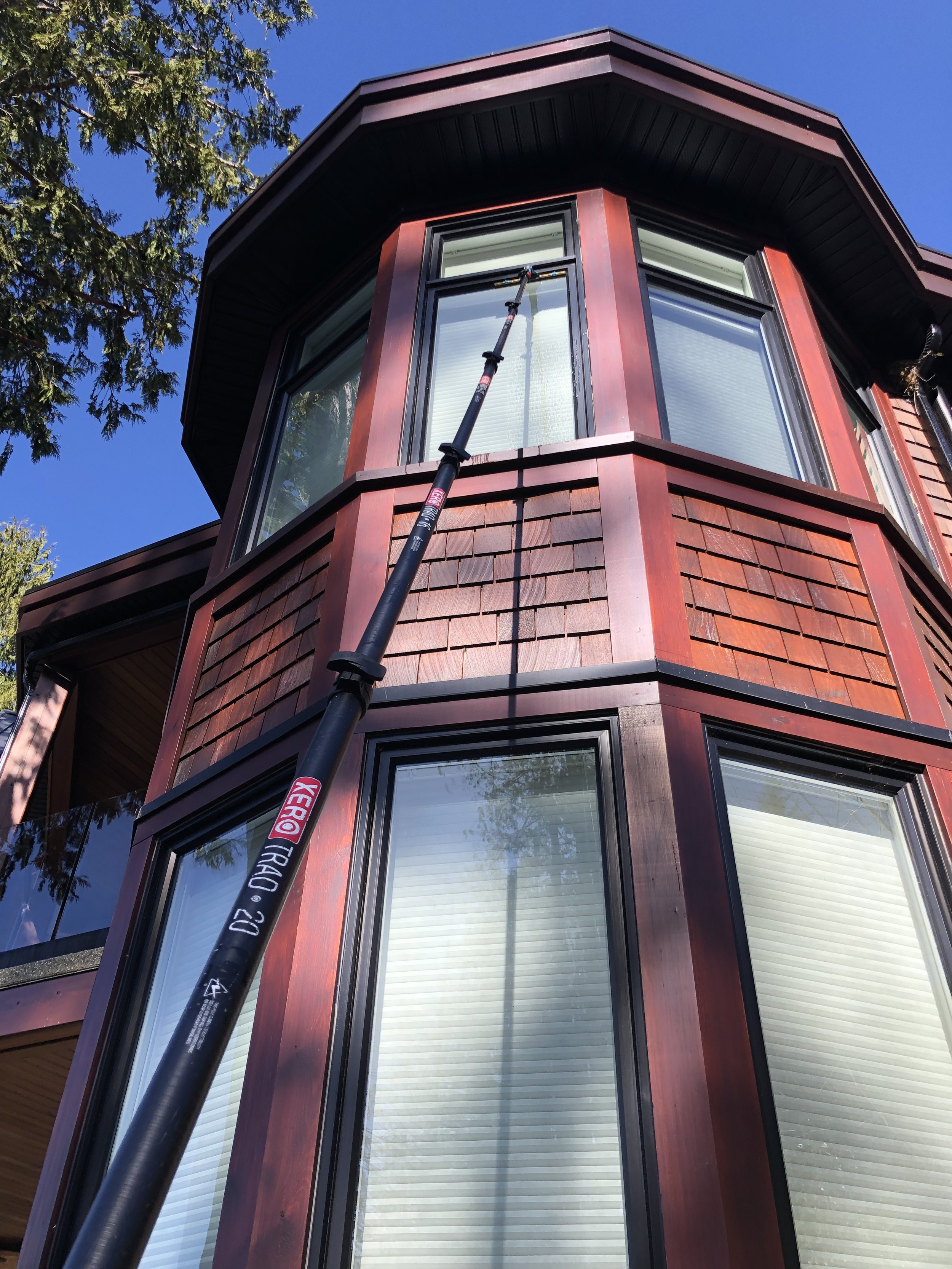 A house with large black-framed windows and brown wooden siding, with a window cleaning pole extended from ground level, reaching the upper window, against a clear blue sky in daytime.