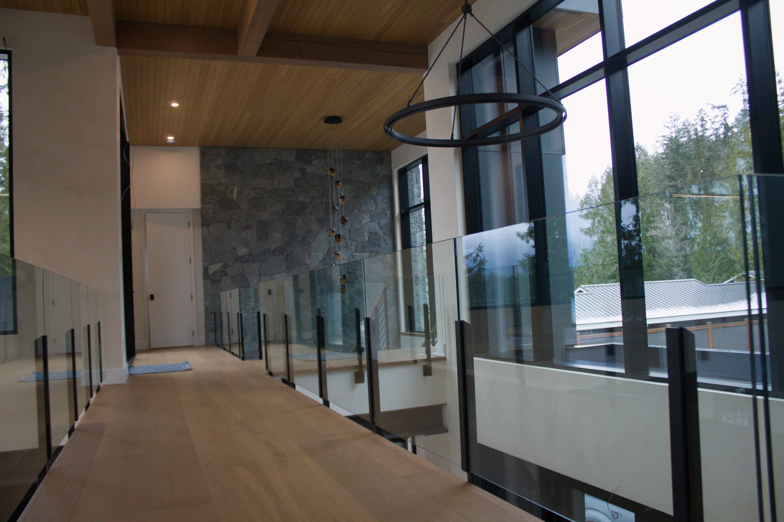 Modern interior with large floor-to-ceiling windows, wooden ceiling, stone accent wall, glass railing, and circular lighting fixture.