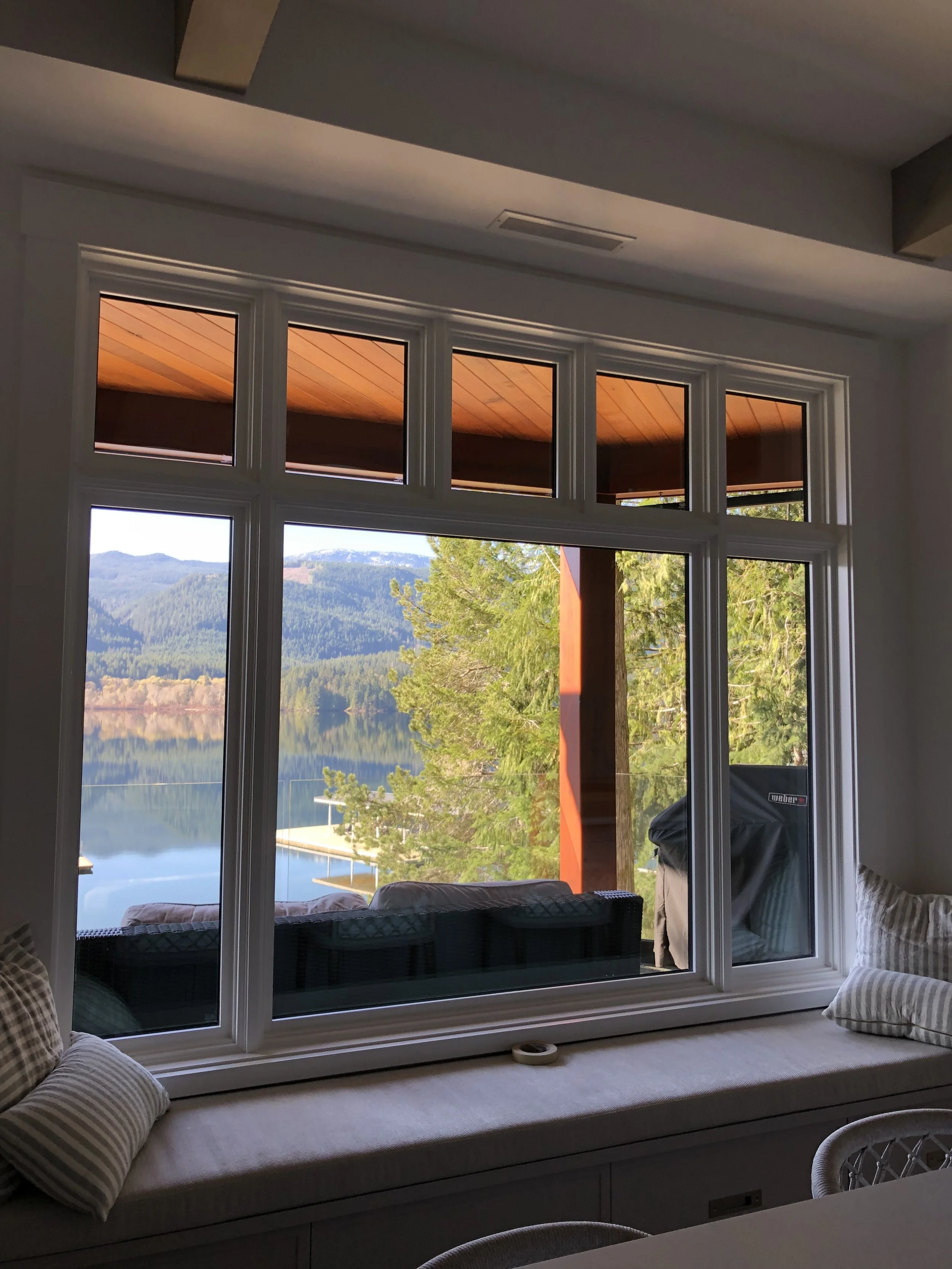 Interior view of a room with a large window overlooking a lake, trees, and mountains. The window has a cushioned window seat with striped pillows, and part of a patio with a grill is visible outside.