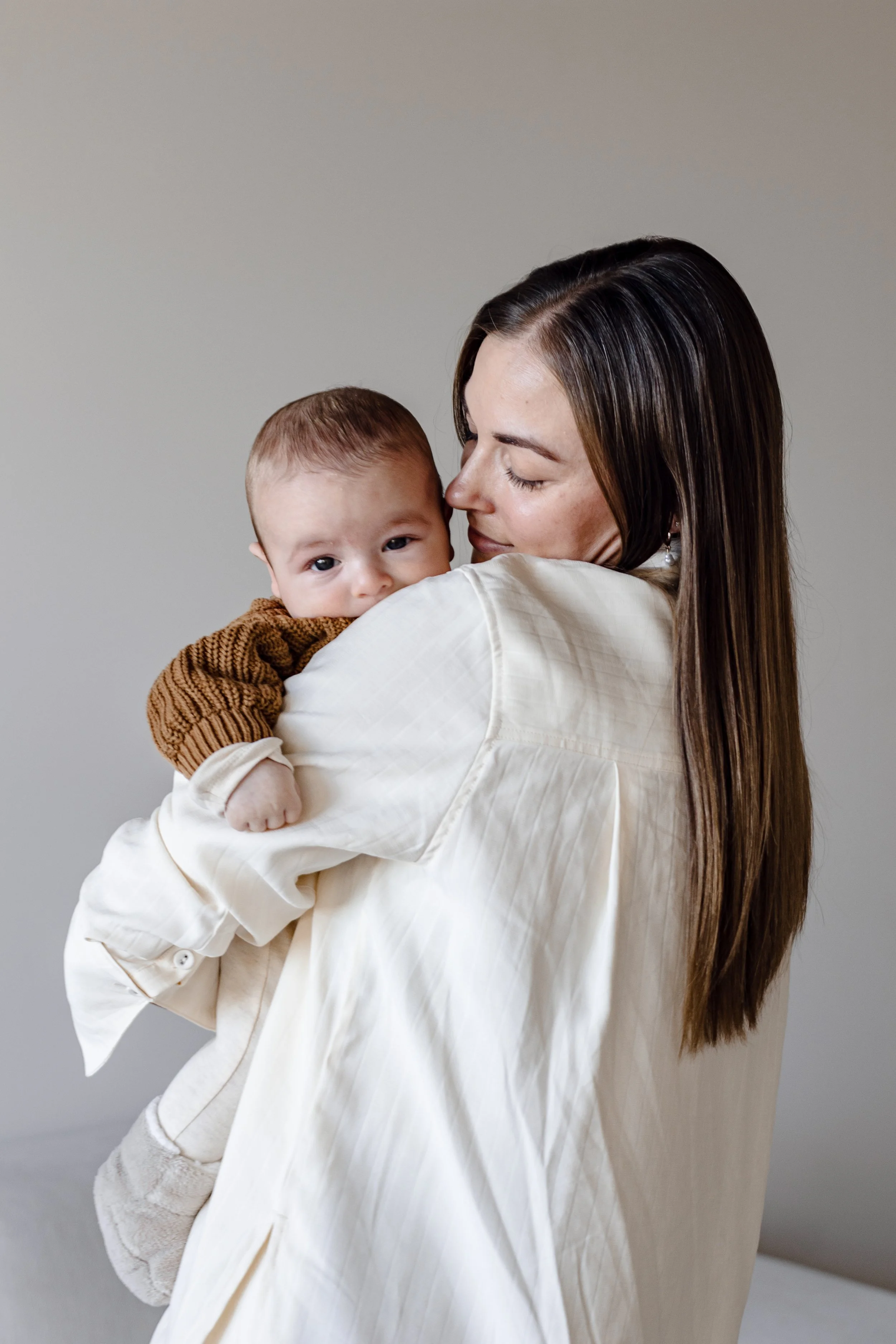 A woman with long brown hair holding a baby, who is looking at the camera, in front of a plain gray background.