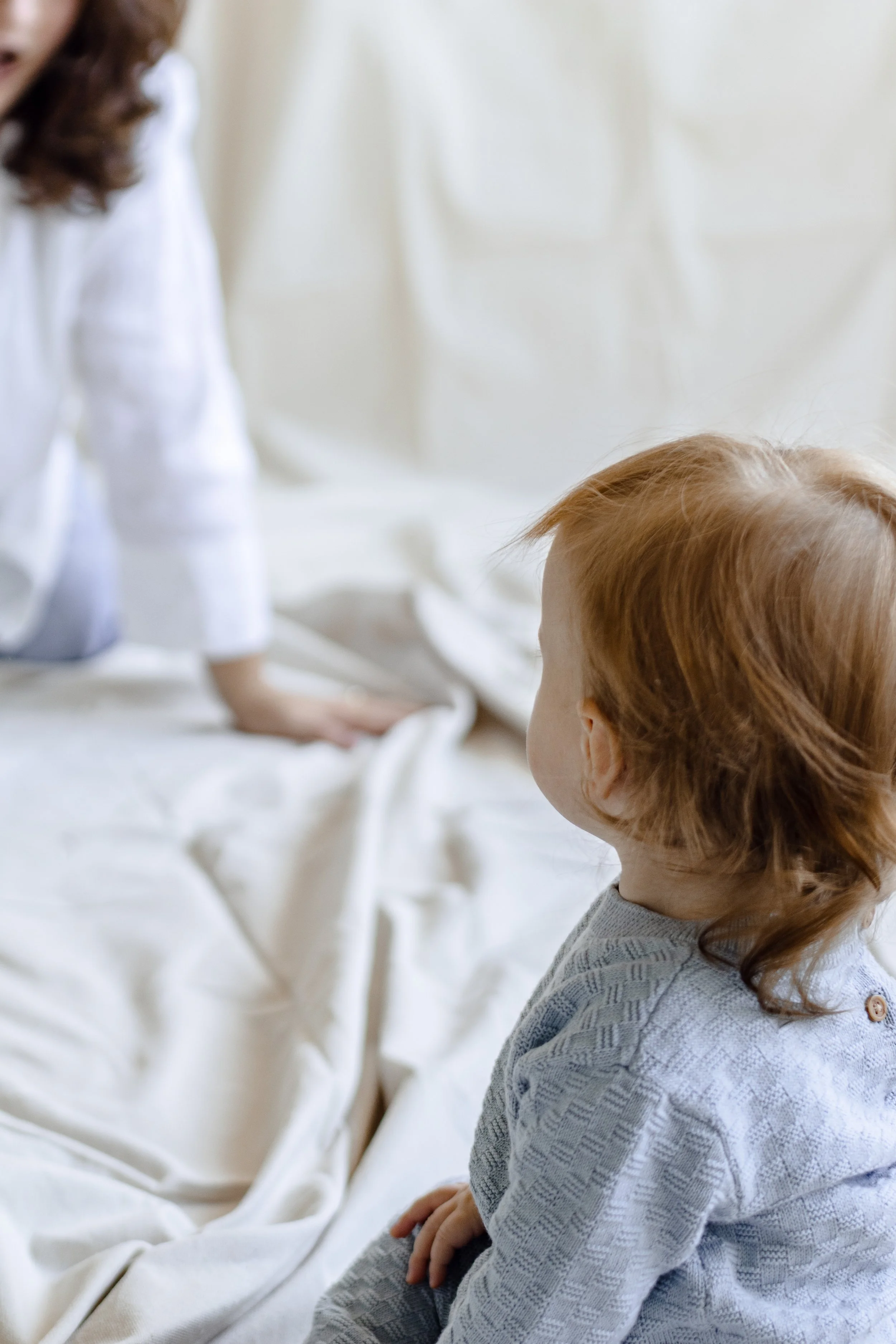 A young child with red hair sitting on a bed, looking at a woman who is kneeling on the bed and reaching out with her hand.