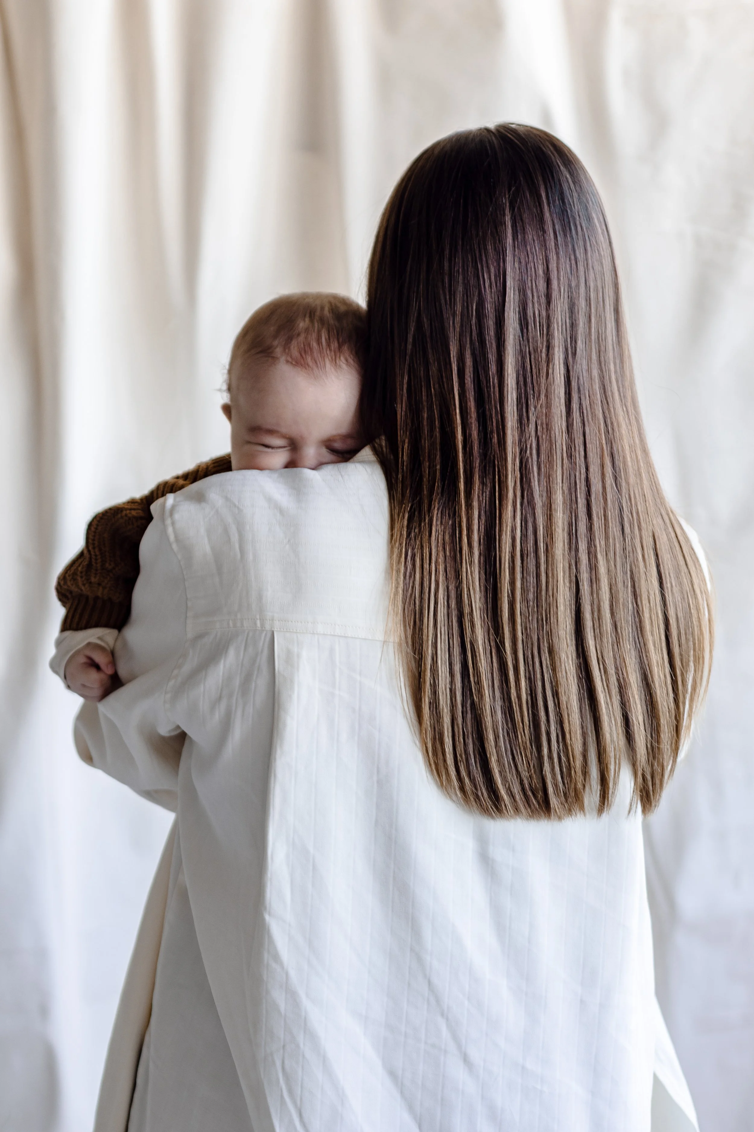 A woman with long brown hair holding a smiling young child with short blonde hair, facing away from the camera, against a white curtain background.