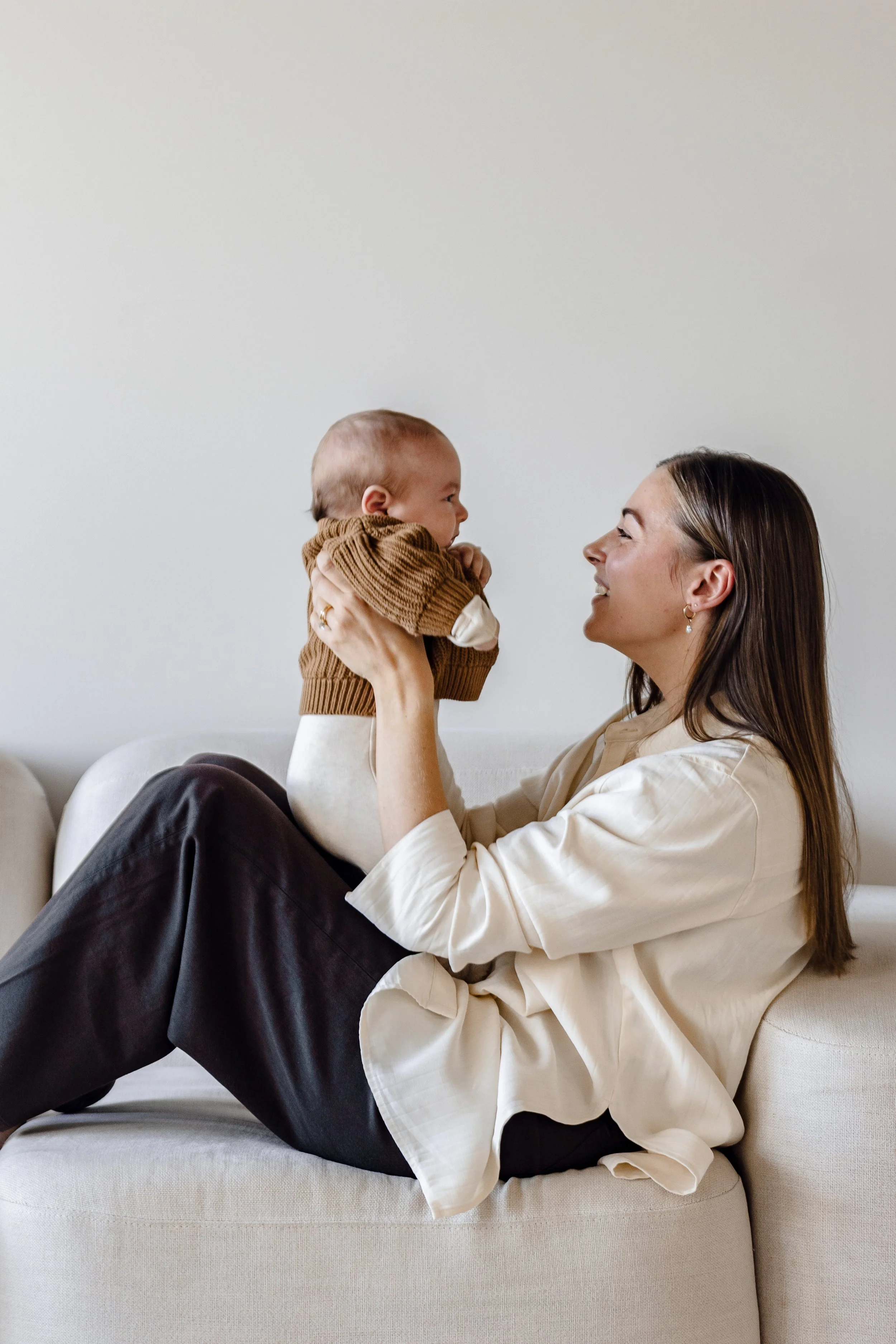 A woman sitting on a white couch, holding a baby in her hands, smiling and facing each other.