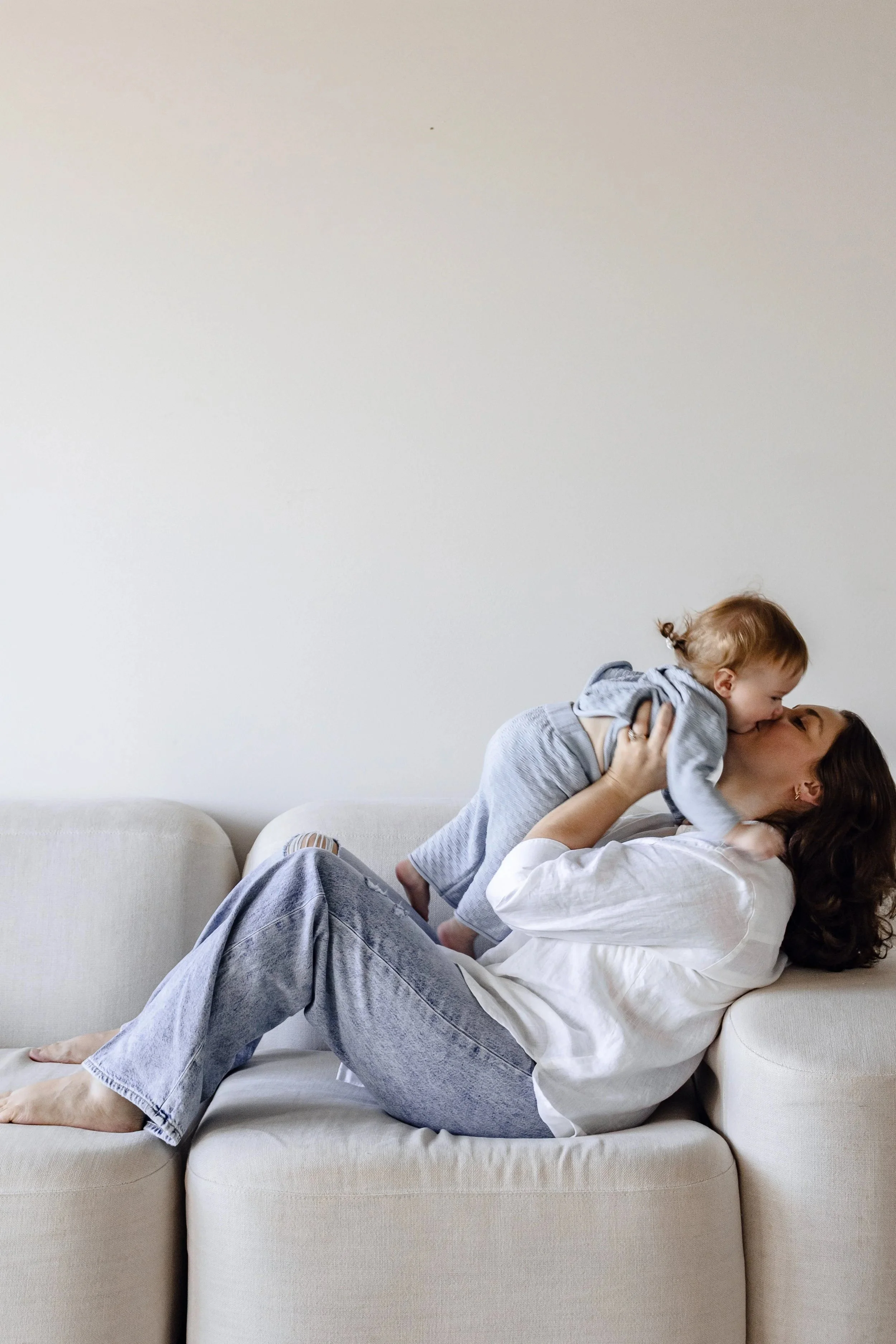 A woman lying on a beige couch, holding a young child up in the air and kissing him.