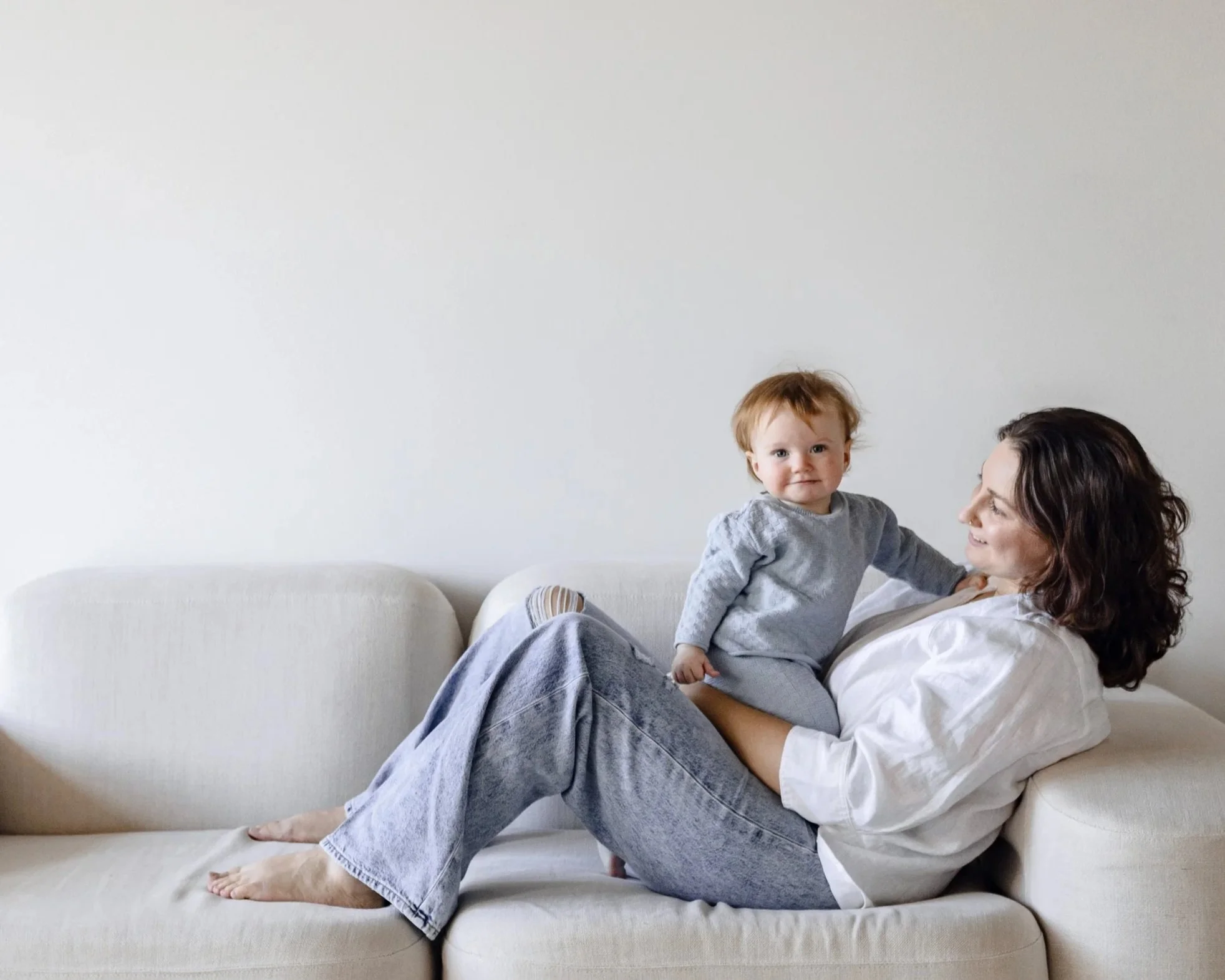 A woman with dark, wavy hair lying on a beige sofa holding a toddler with light brown hair in a gray outfit.