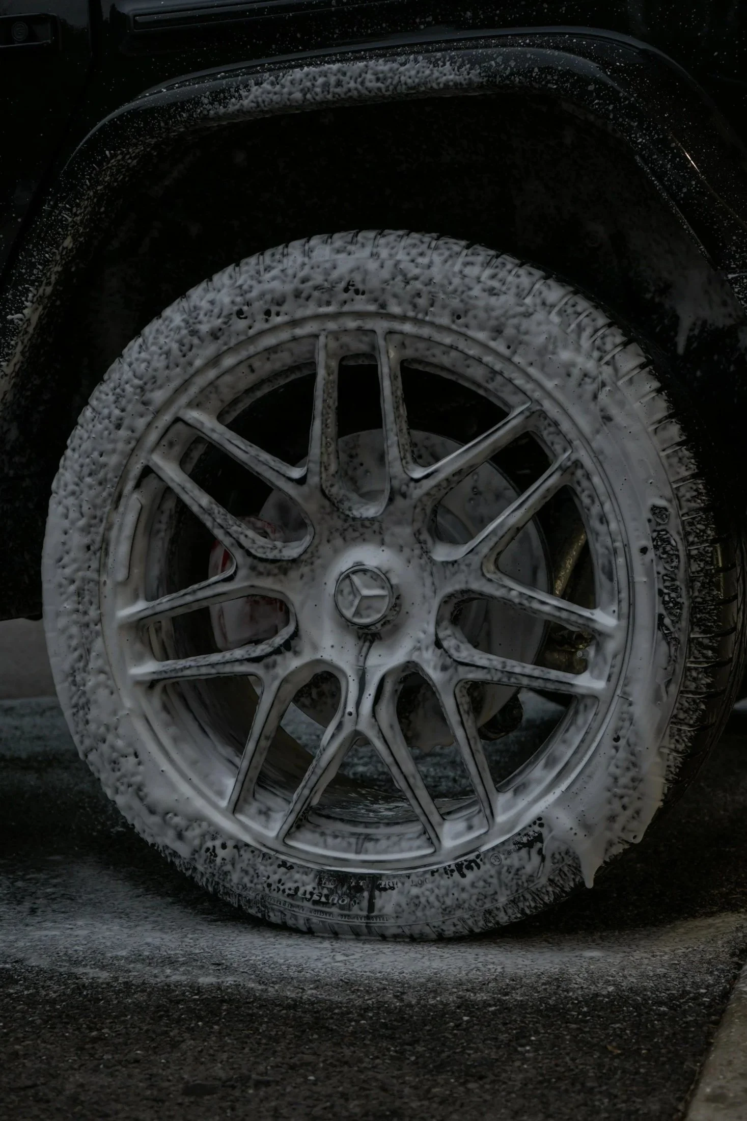 Close-up of a car wheel covered in foam soap, with foam dripping down the side of the tire and rim, on a dark surface.