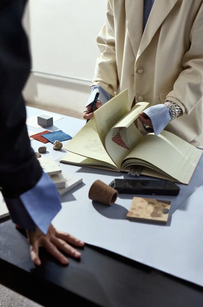 A person in a beige coat and blue shirt is showing a large open book with sketches to another person, whose hand is resting on the table. The table has fabric samples, small objects, and design tools.