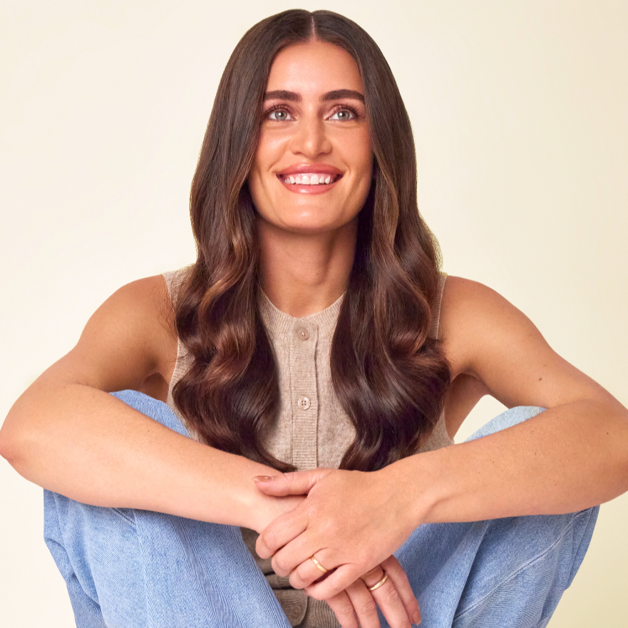 A smiling young woman with long, wavy brown hair, wearing a sleeveless beige top and sitting with her arms wrapped around her knees, positioned against a plain light-colored background.