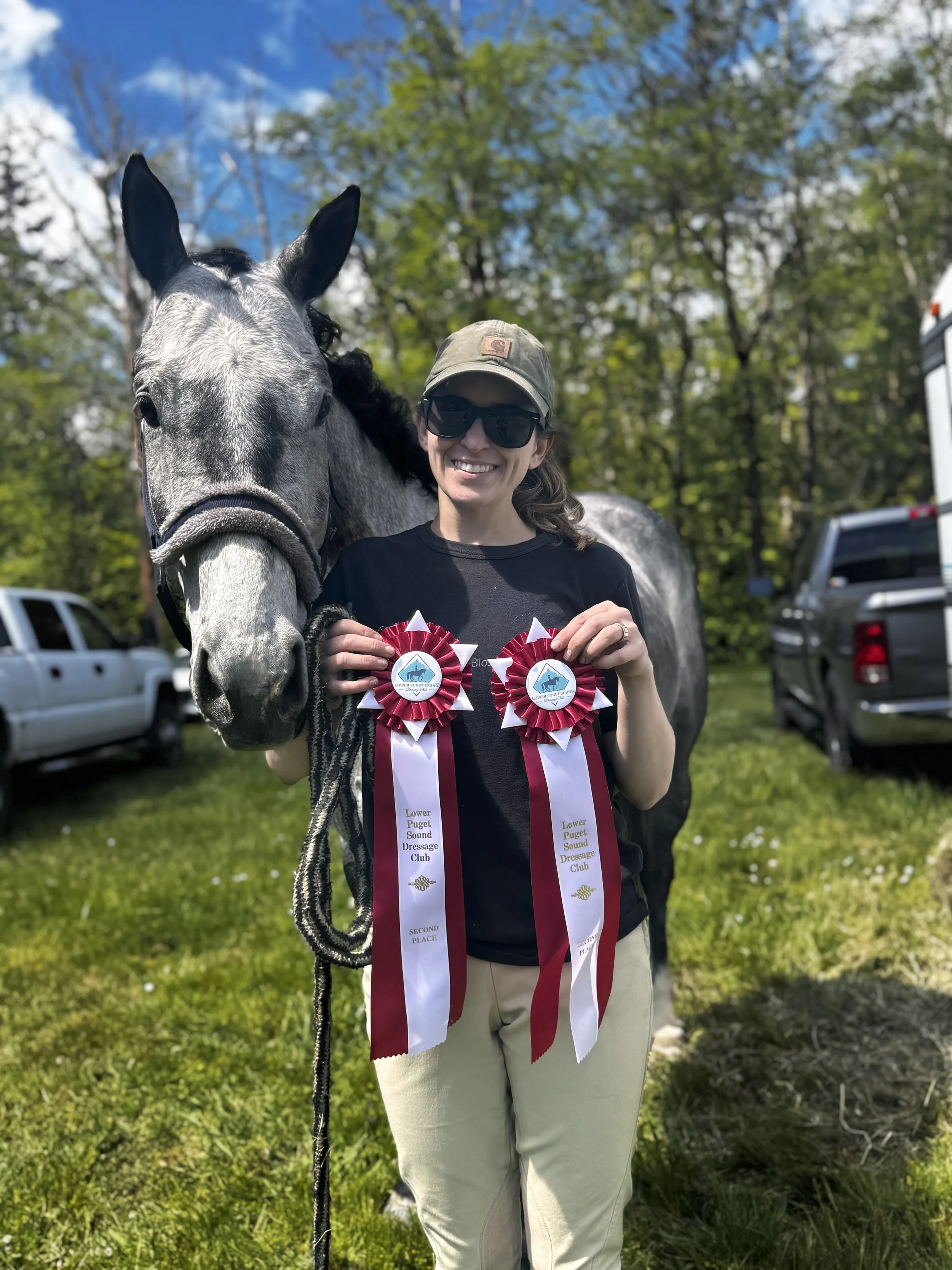 A woman wearing sunglasses, a cap, and a black shirt is smiling while holding two rosettes from the Lower Puget Sound Dressage Club, standing outdoors with a gray horse. The background features green trees, cars, and a bright blue sky with some clouds.