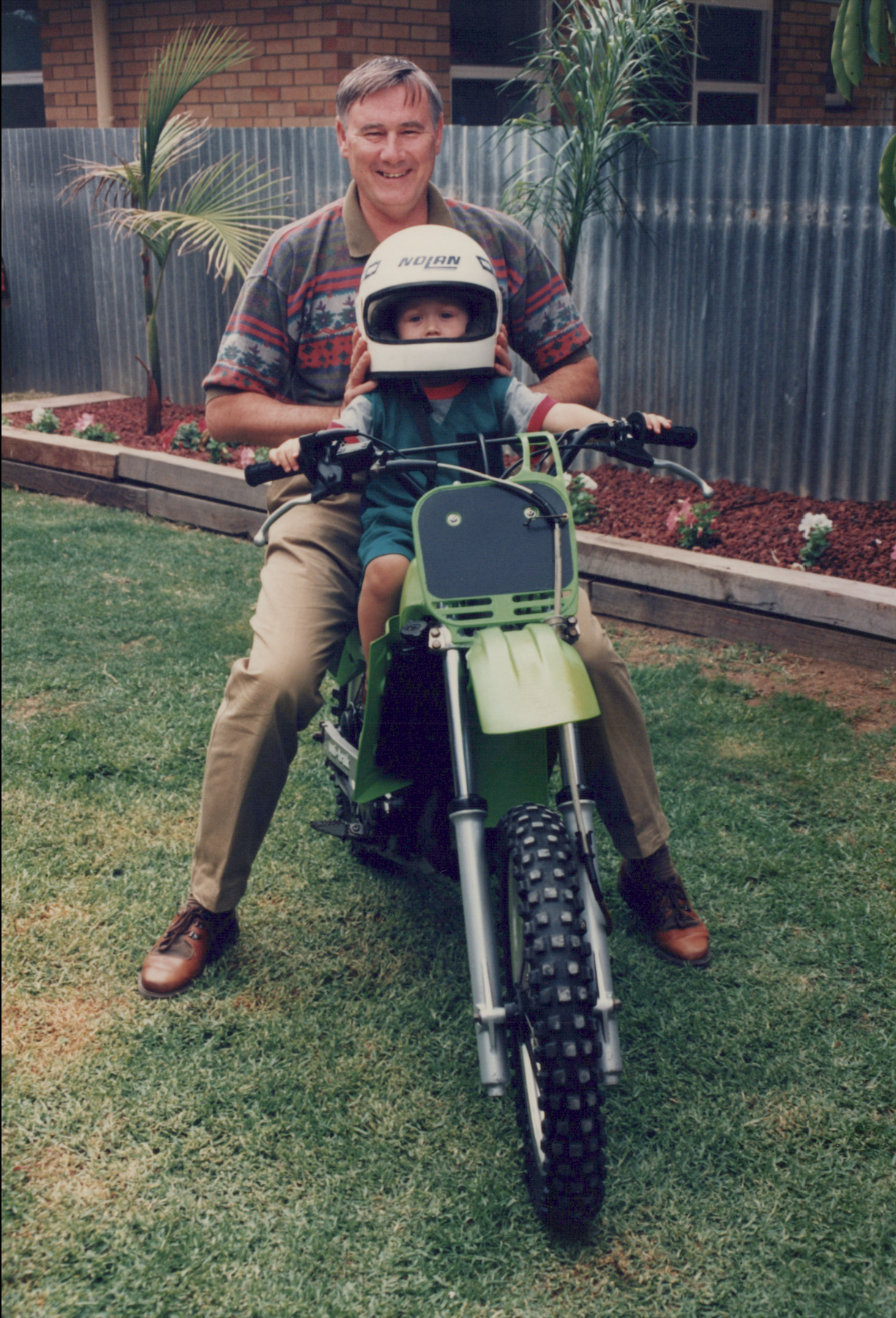 A man and a child on a dirt bike in a backyard with grass, palm trees, and a corrugated metal fence.