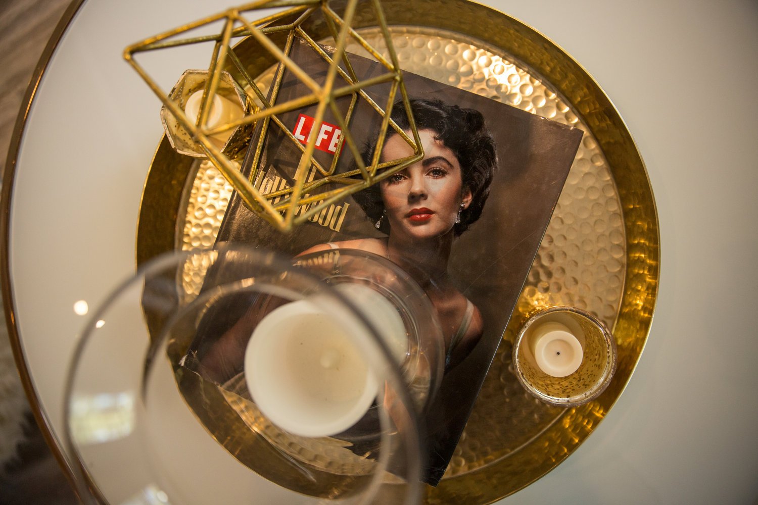 Top-down view of a gold tray with a candle, a magazine featuring Elizabeth Taylor with dark curly hair and red lipstick, a decorative gold geometric object, and a smaller candleholder with a candle inside.