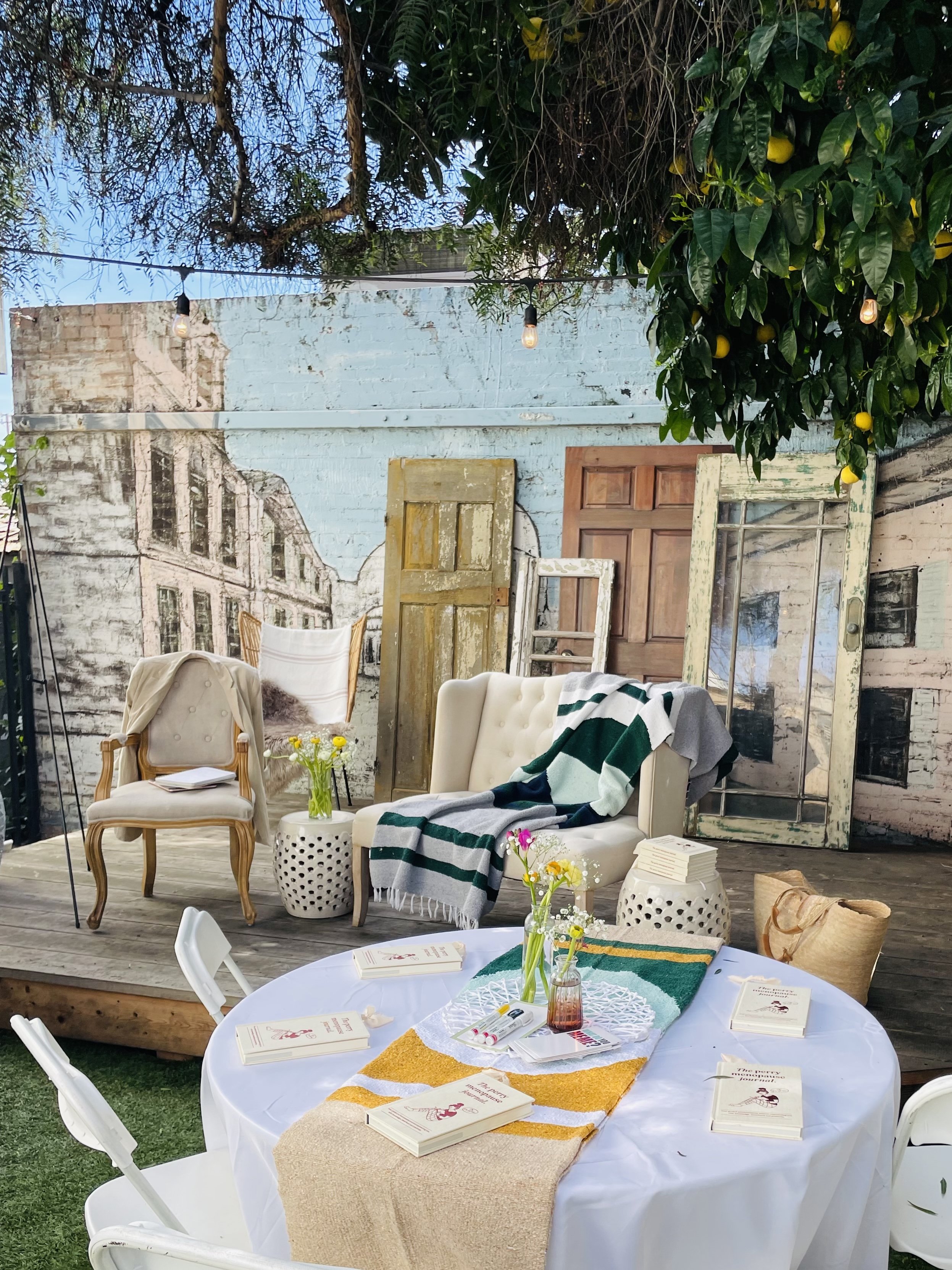 A decorated outdoor space with a white round table on grass, surrounded by white chairs. The table has a yellow and white runner, a burlap cloth, books, and small white cards. Behind the table, there's a patio area with vintage doors and window frame