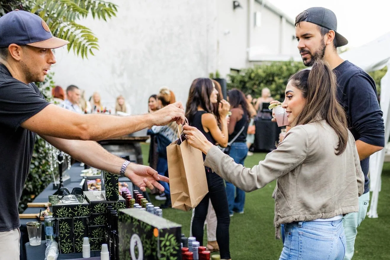 A young woman in a beige jacket and blue jeans buying olive oil and receives bag from a man at an outdoor event, with other people socializing in the background.