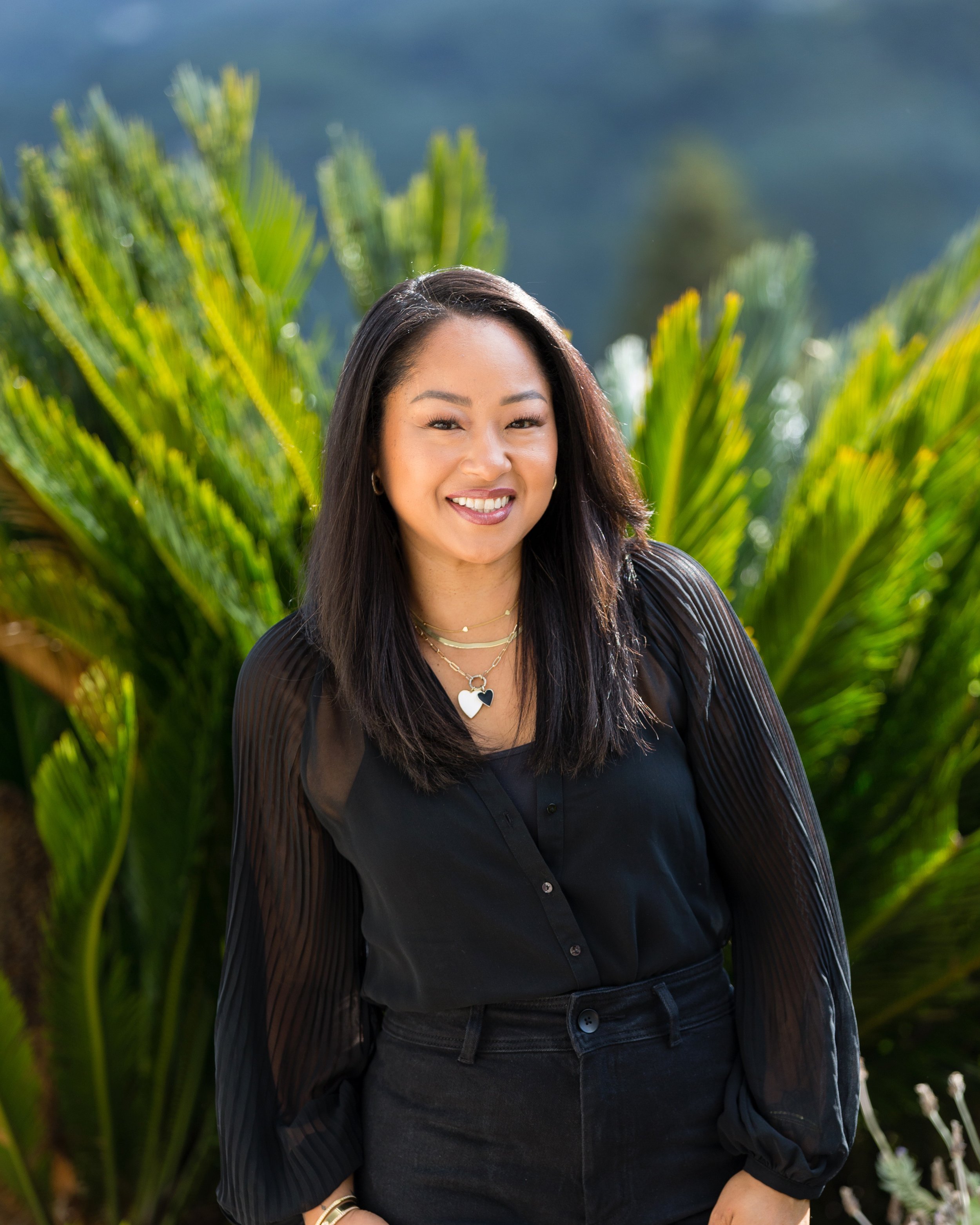 A woman with long dark hair, smiling, wearing a black top, layered necklaces, and gold bracelets, standing outdoors in front of lush green plants.