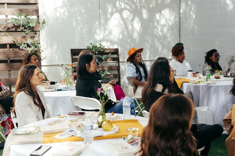Group of women sitting at tables outdoors during a daytime event, some with drinks and notes, some taking photos, with plants and trees in the background.