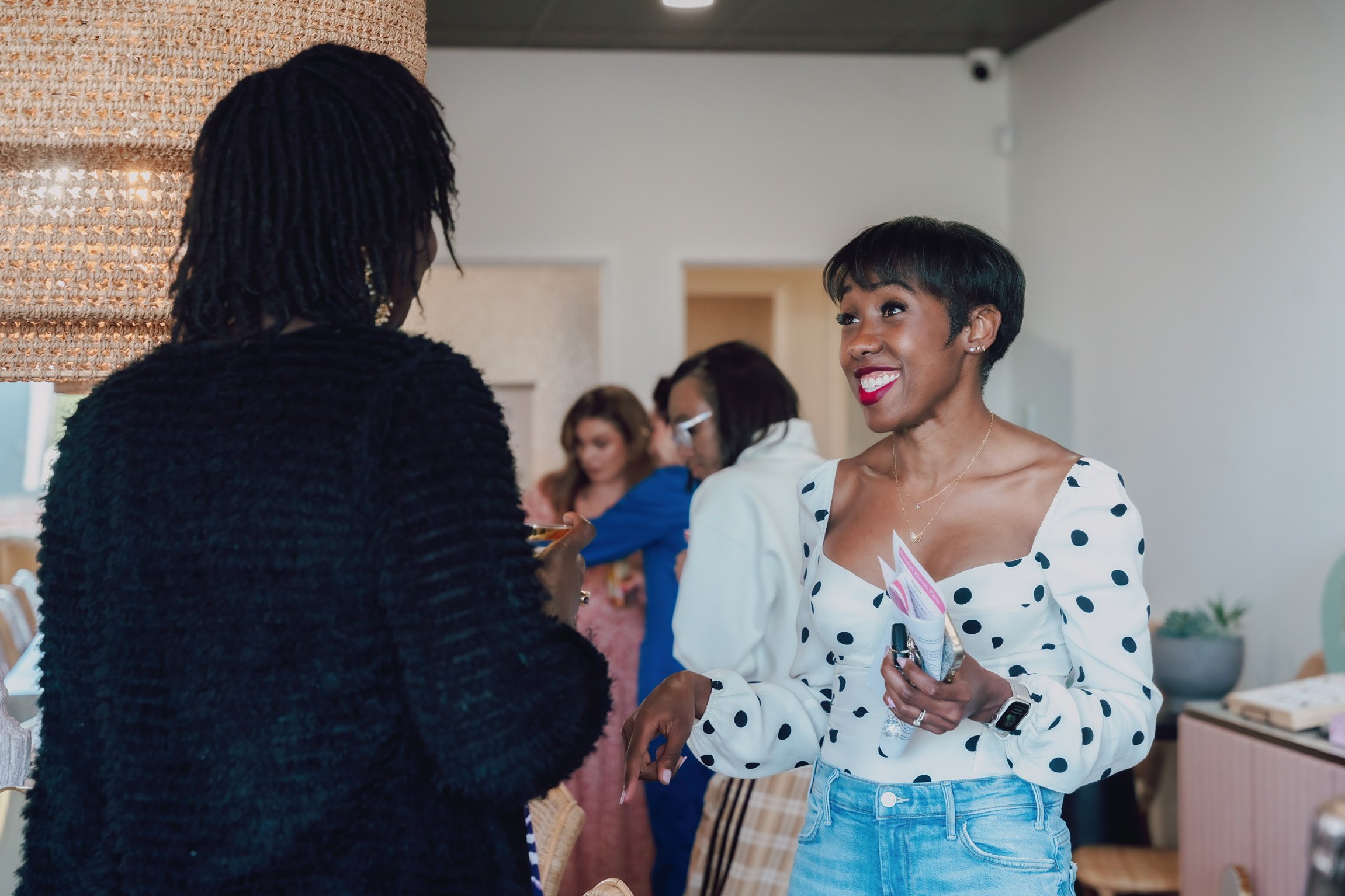 A woman with short black hair, wearing a white shirt with black polka dots, is smiling and talking to another woman with locs, who is dressed in black. Other people are visible in the background.