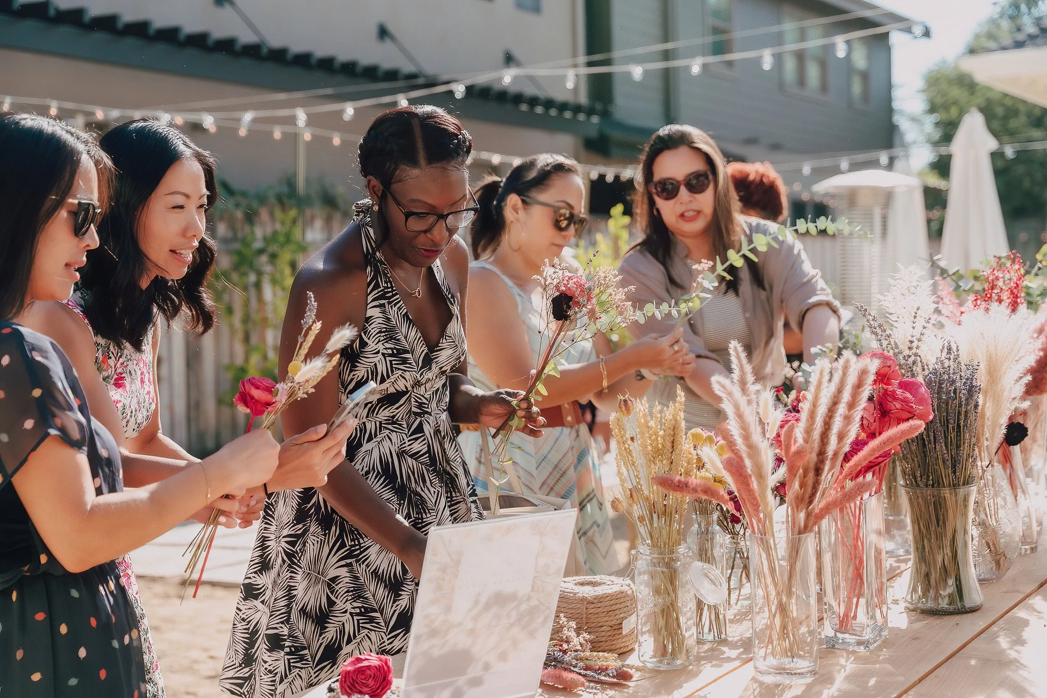 A group of women at an outdoor event, standing behind a table decorated with various dried flowers, creating holding flower bouquets and looking down at the display.