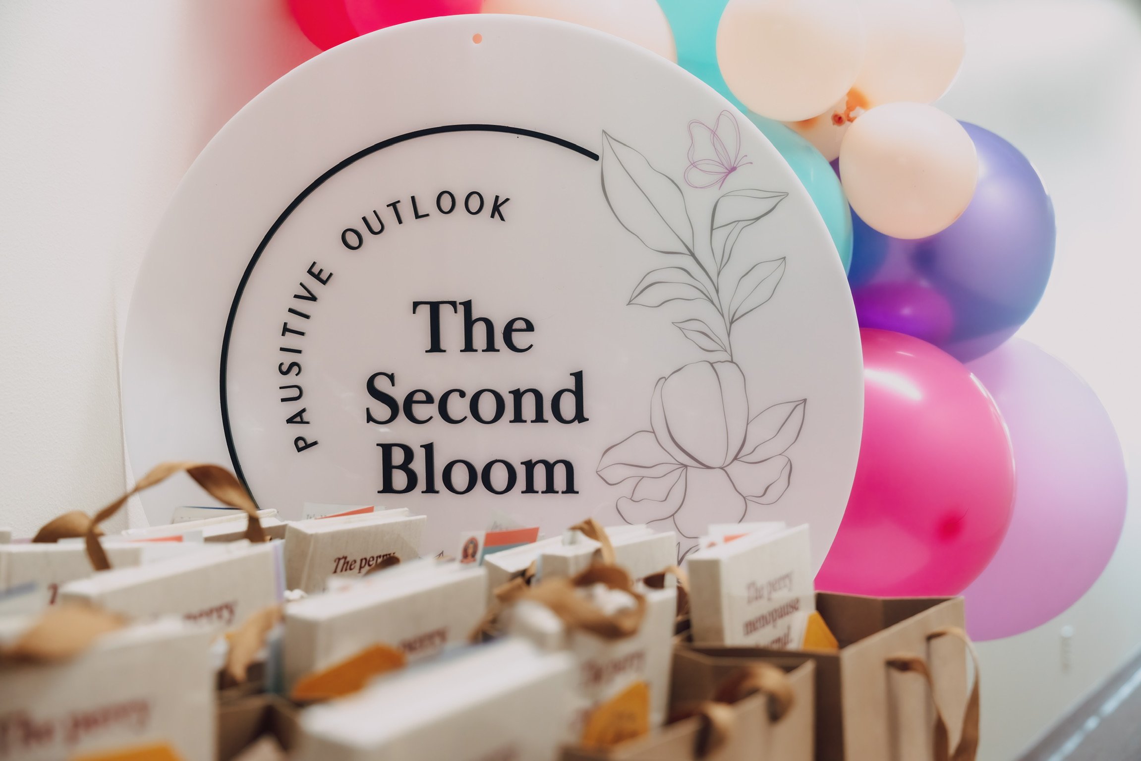 Decorative sign with the words 'The Second Bloom' and floral line drawing, surrounded by colorful balloons in pink, purple, and blue, and small bags or gift items in the foreground.